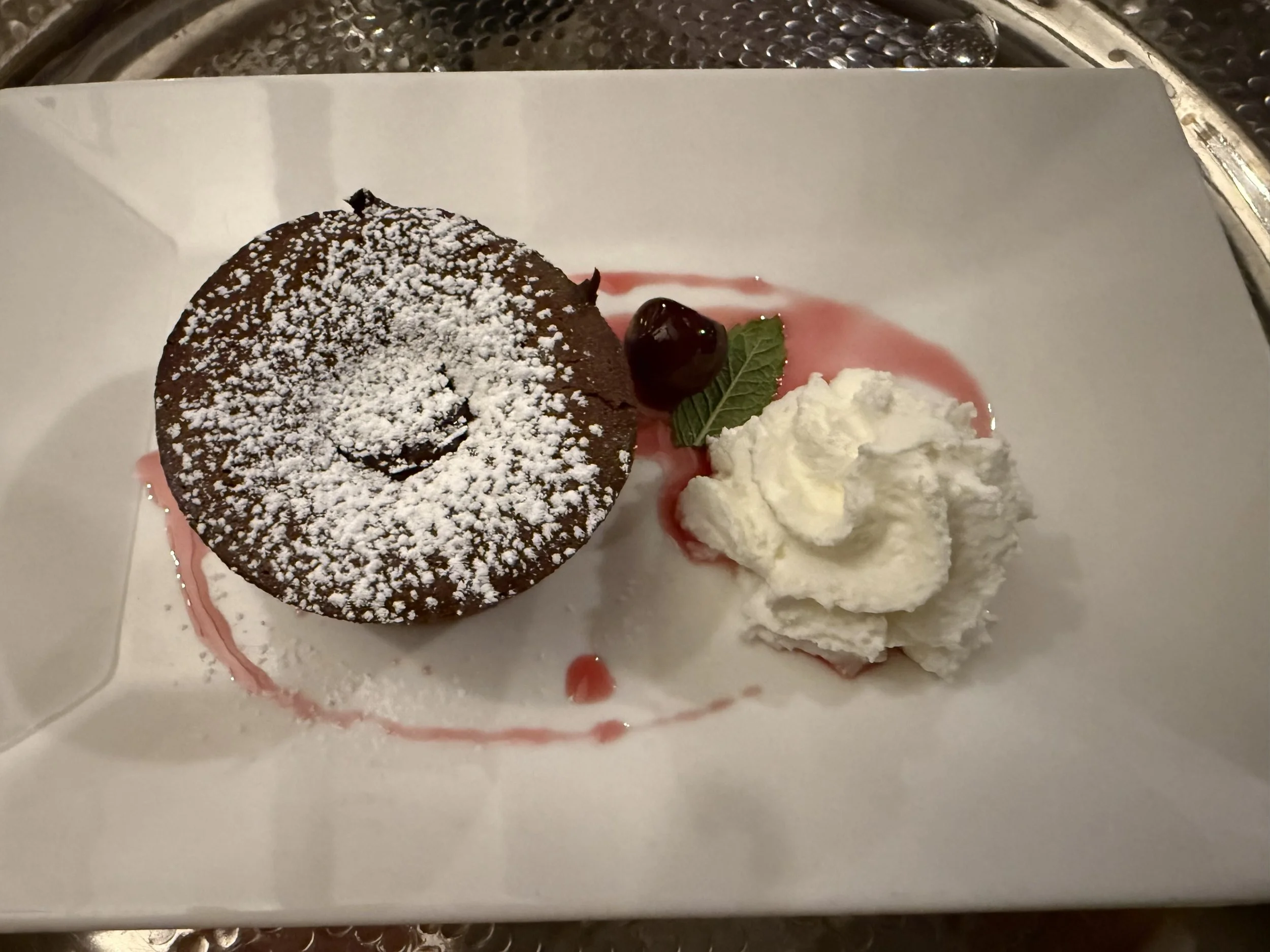 A chocolate lava cake dusted with powdered sugar, served with a dollop of whipped cream, a cherry, and a mint leaf on a white plate with a pink sauce drizzle.