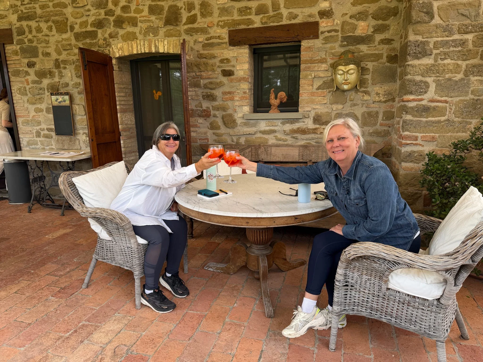 Two women sitting at an outdoor table raising their glasses together for a toast, with a rustic brick building wall behind them.