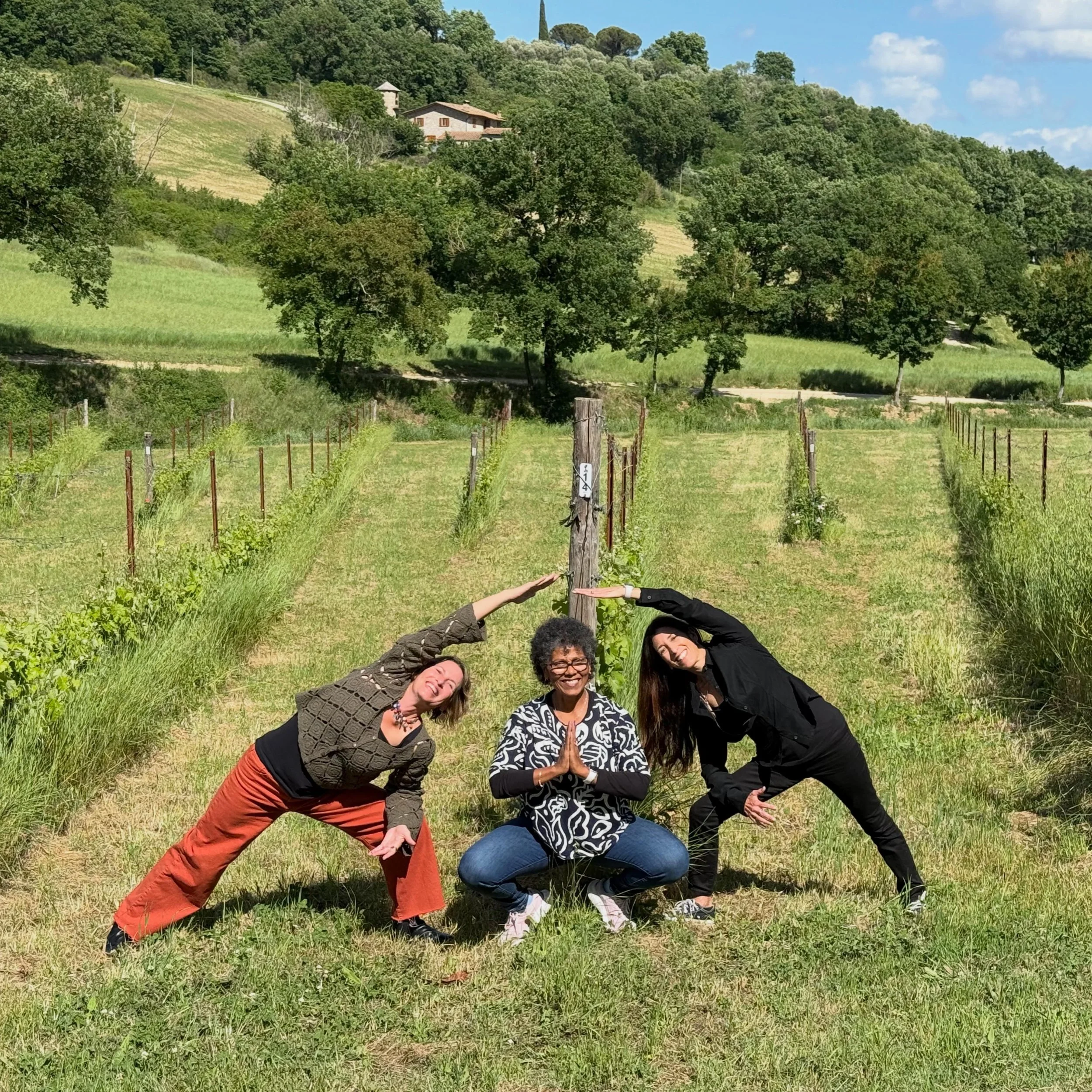 A group of five diverse women practicing yoga outdoors in a green vineyard with rolling hills and houses in the background on a sunny day.