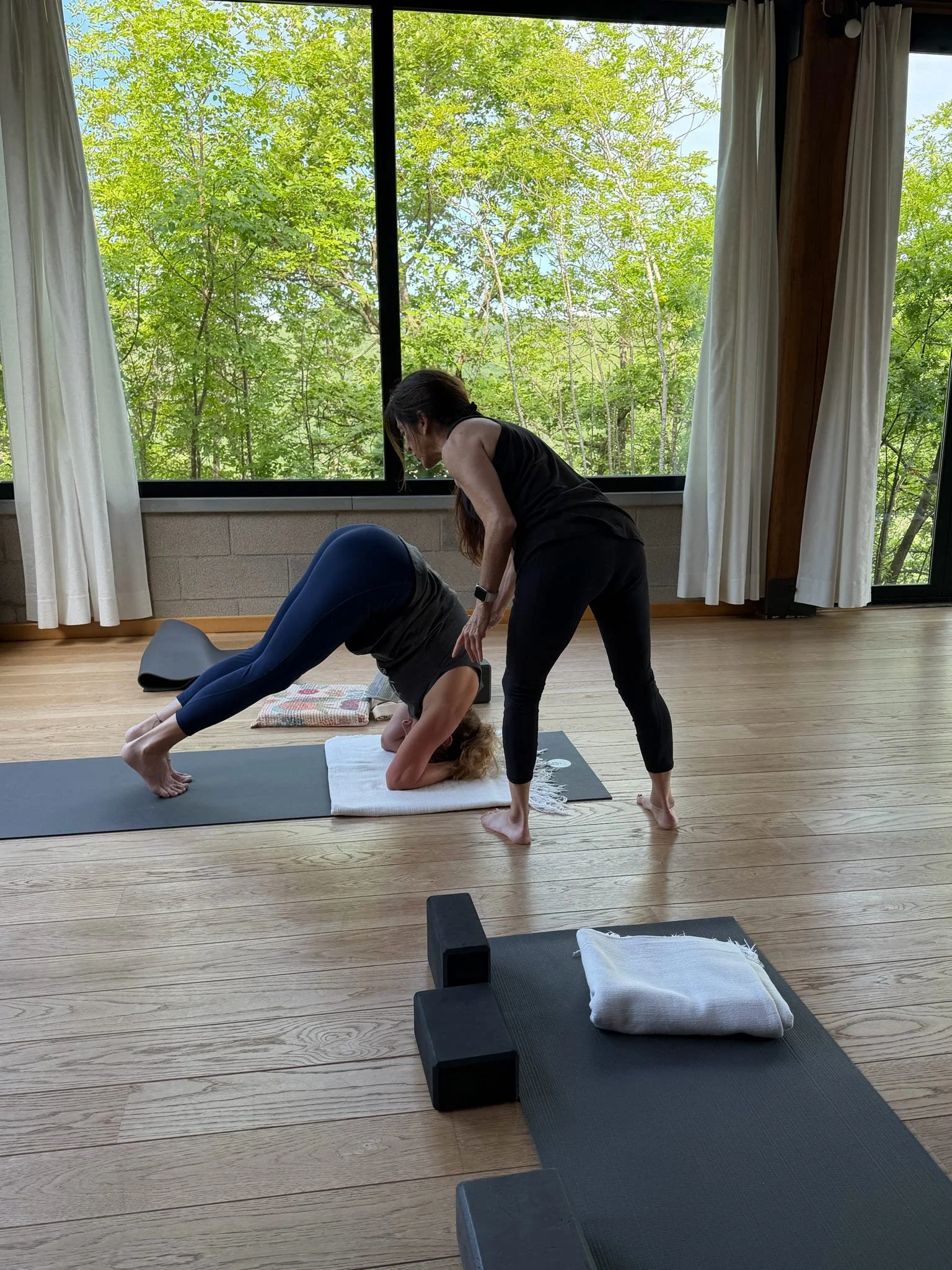 A woman in black leggings and a black top practicing yoga inside a room with wooden floors and large windows. She is in a forward bend position with her head between her legs, and another woman is assisting her by holding her hips. Yoga mats, towels,