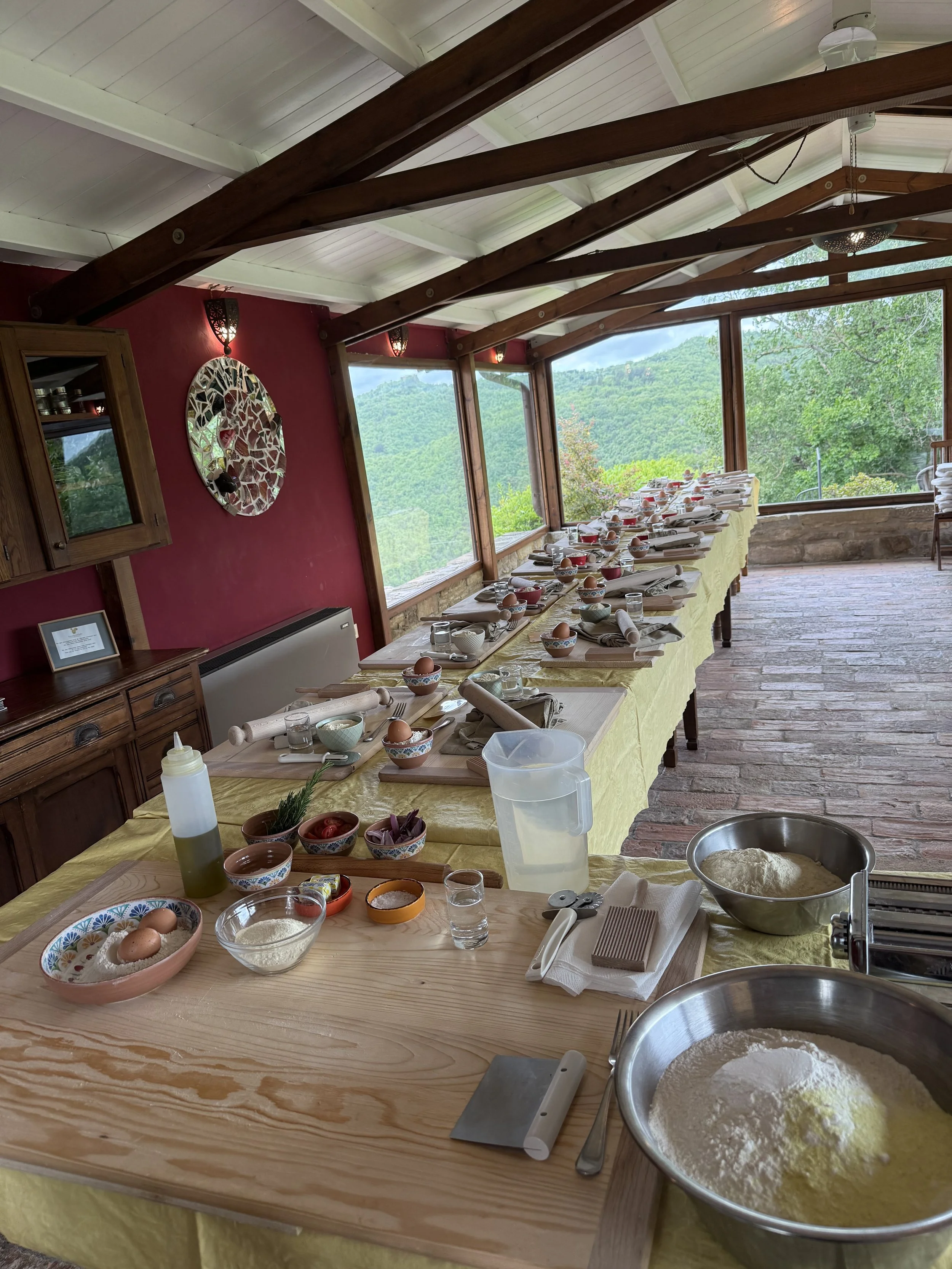 A long table set for a cooking or baking activity, with bowls of flour, eggs, measuring cups, rolling pins, and other baking tools in a bright room with large windows overlooking green hills.