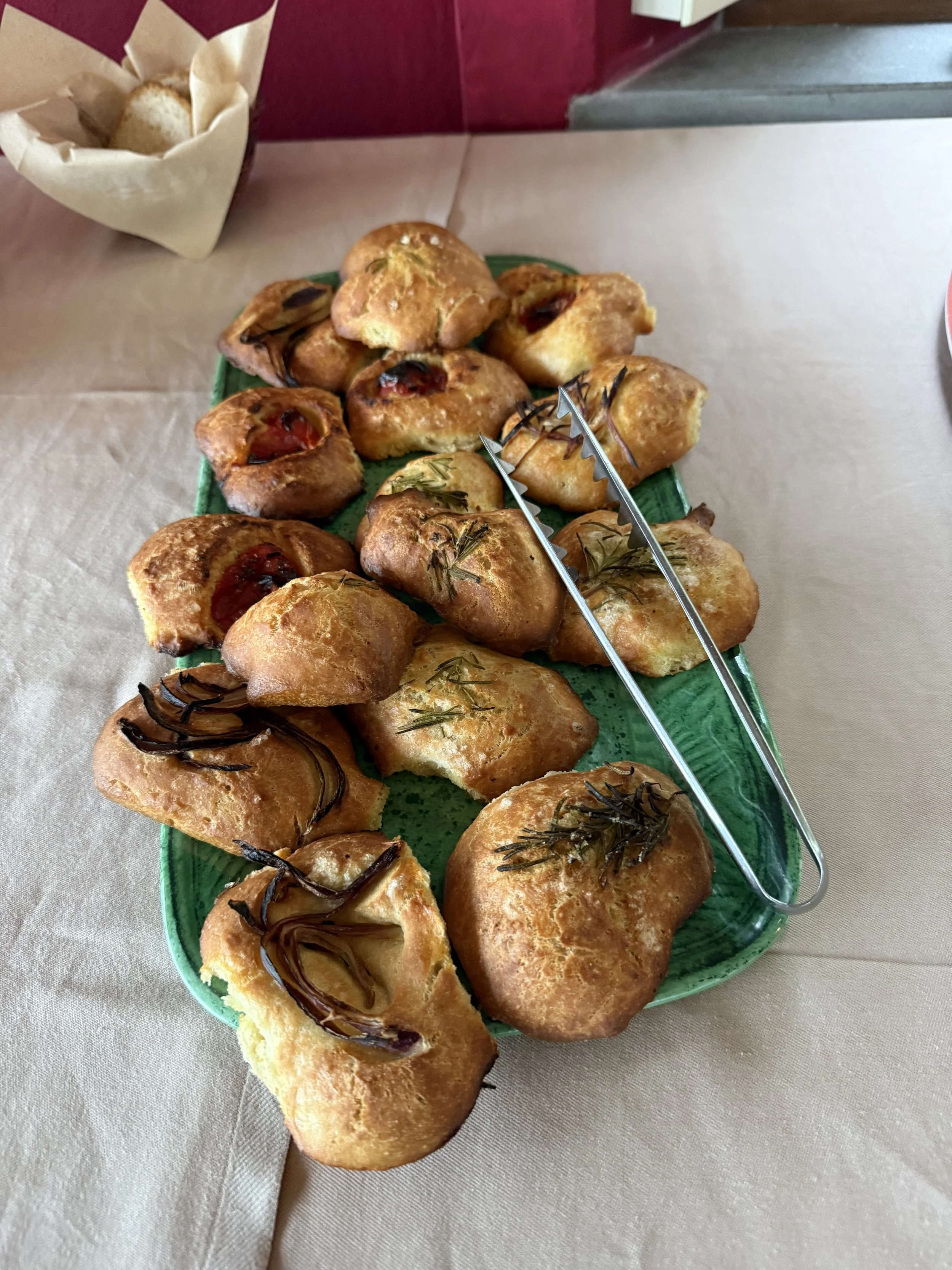 A green platter with assorted baked bread rolls topped with herbs and chocolate, placed on a beige tablecloth. Tongs rest on the platter. In the background, a small paper bag with bread rolls is visible.