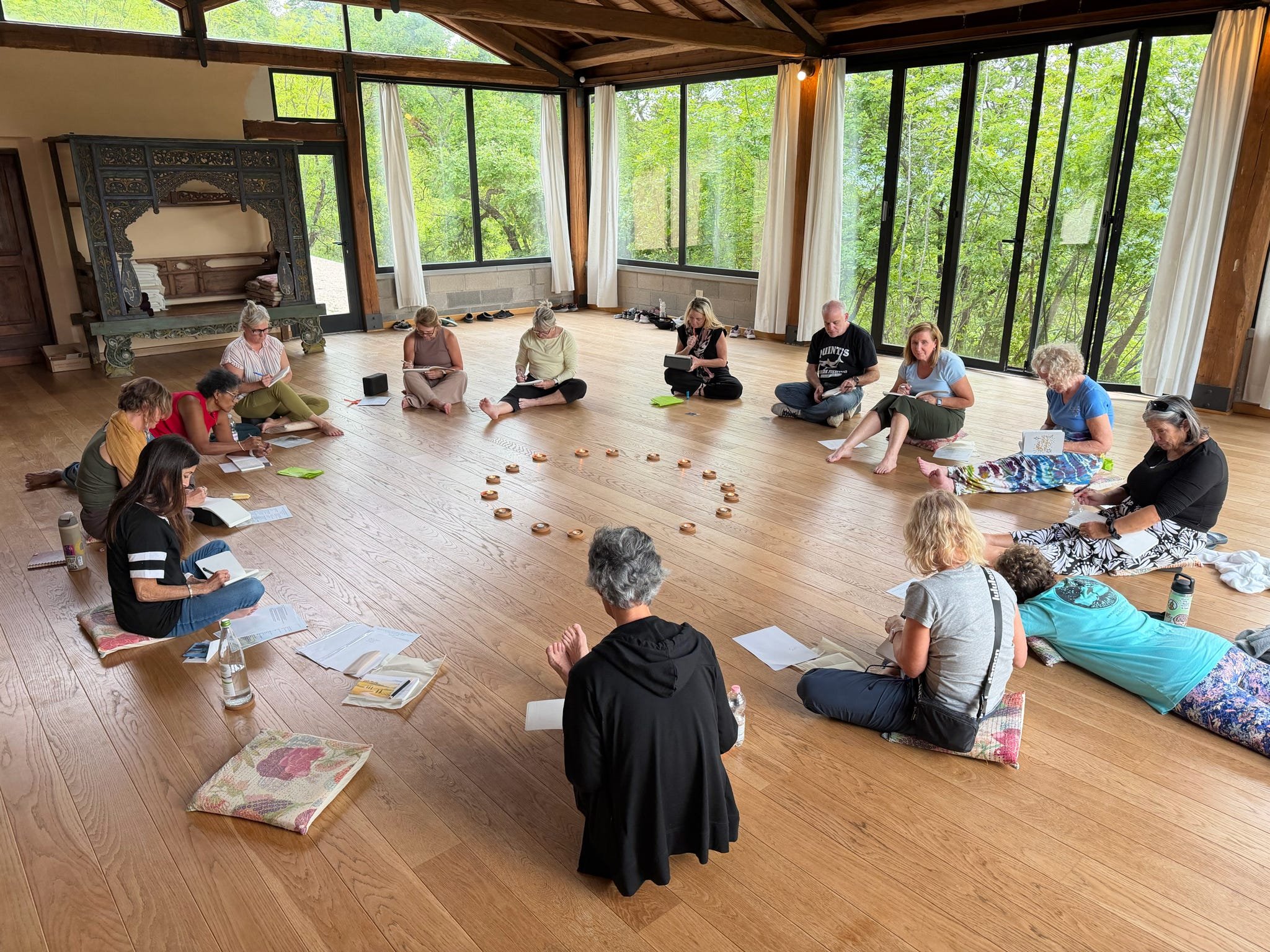 A group of people sitting cross-legged on cushions and the wooden floor of a spacious room with large windows showing green trees outside. They are engaged in a meditation or prayer session, some holding notebooks or papers.