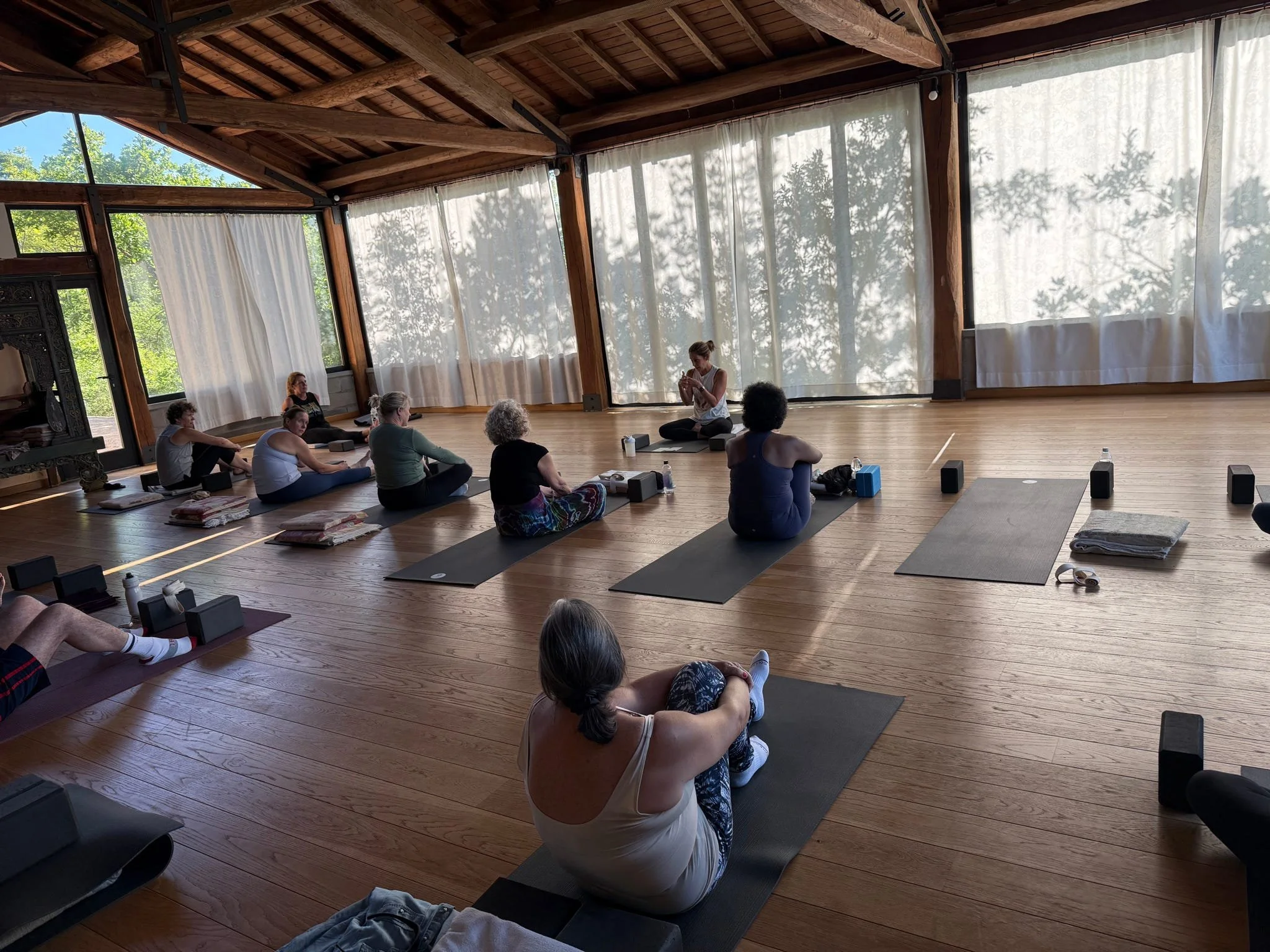 A group of people participating in a yoga or meditation class in a spacious wooden room with large windows and curtains, seated on yoga mats.