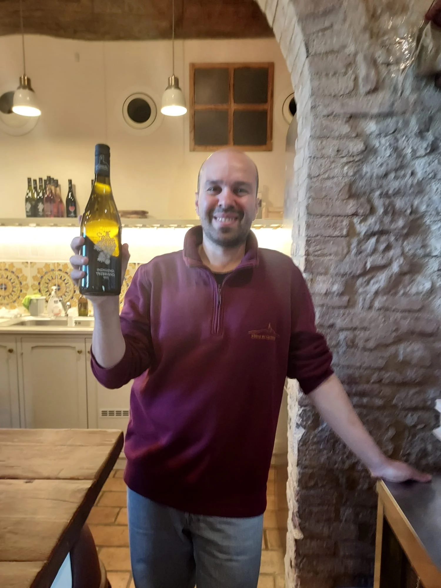A smiling man with a beard holding a beer bottle indoors near a brick wall and a wooden table.