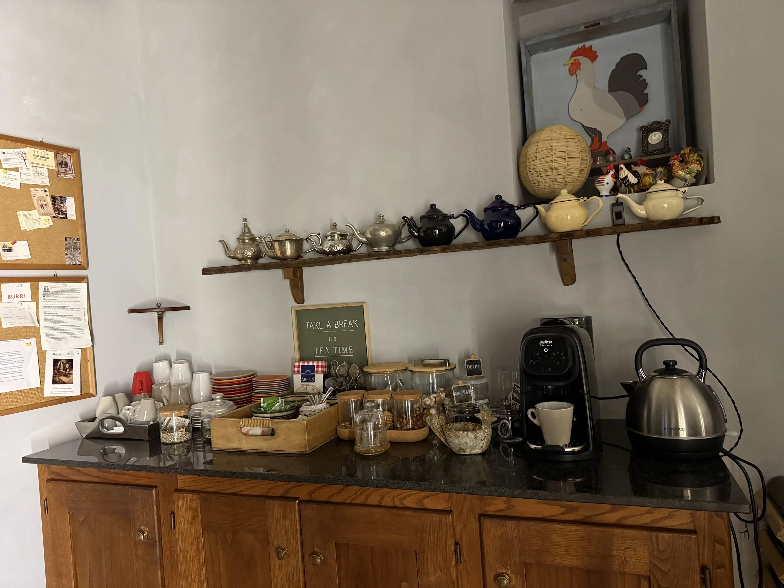 A kitchen countertop with various coffee cups, glasses, and jars. A wooden shelf holds teapots and decorative items. A coffee maker and electric kettle are on the right side. A purple framed sign says 'Take a break, it's tea time.' A bulletin board w