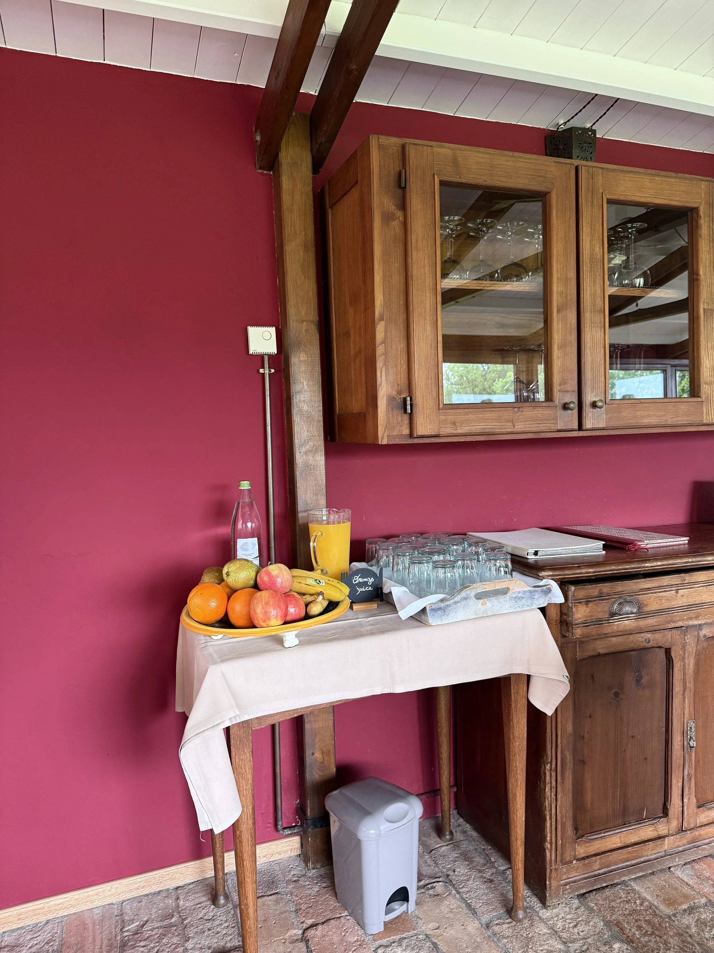 A small table covered with a beige tablecloth holds a bowl of assorted fruits, a bottle of water, a glass of orange juice, and glasses of water. Behind the table, there is a wooden cabinet with glass doors containing wine glasses. The wall behind the