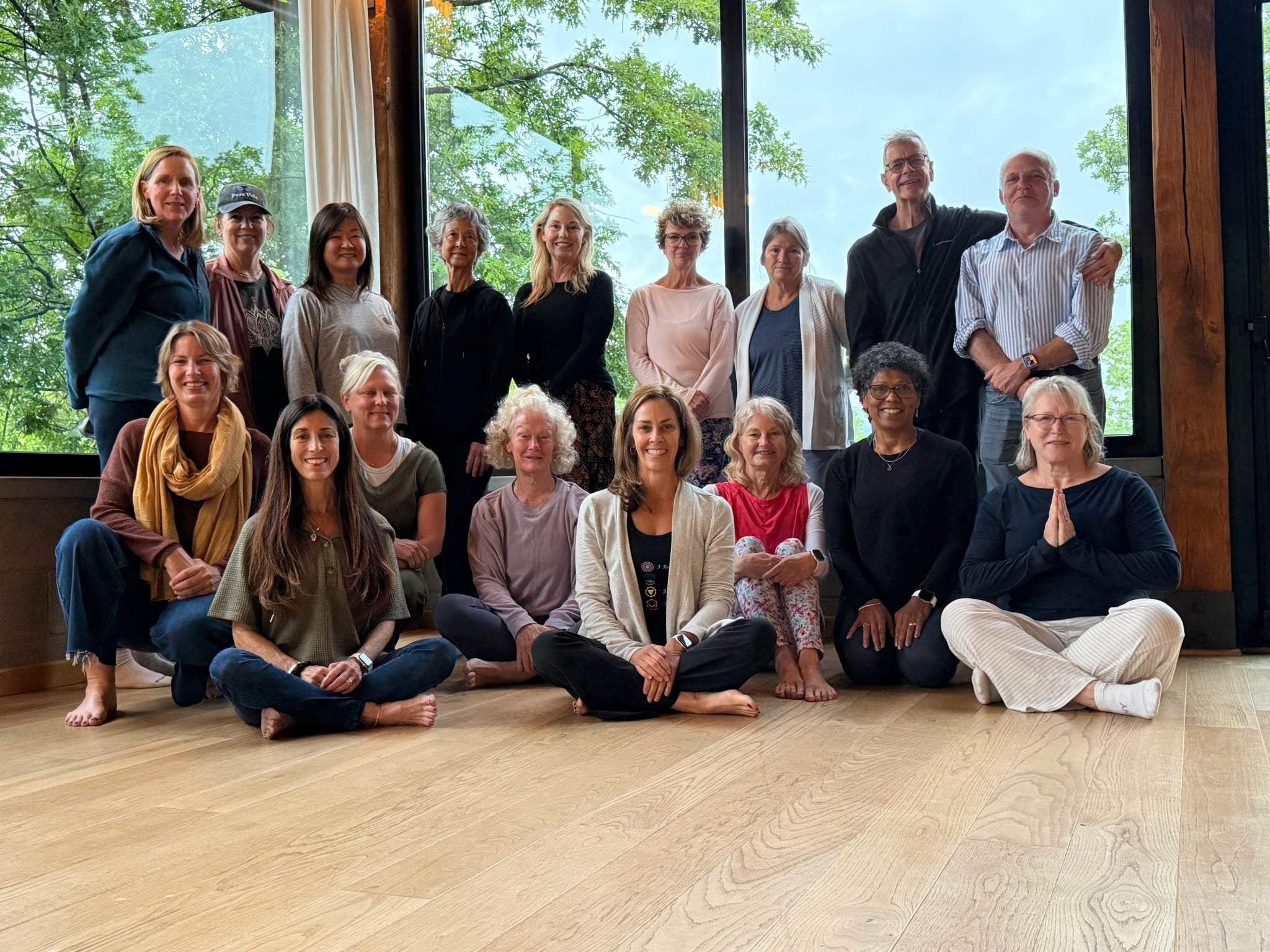 A group of 16 people, mostly women, pose for a photo inside a room with large windows showing green trees outside. Some are sitting on the wooden floor, and others are standing behind them. They are smiling and appear relaxed, with some barefoot and 