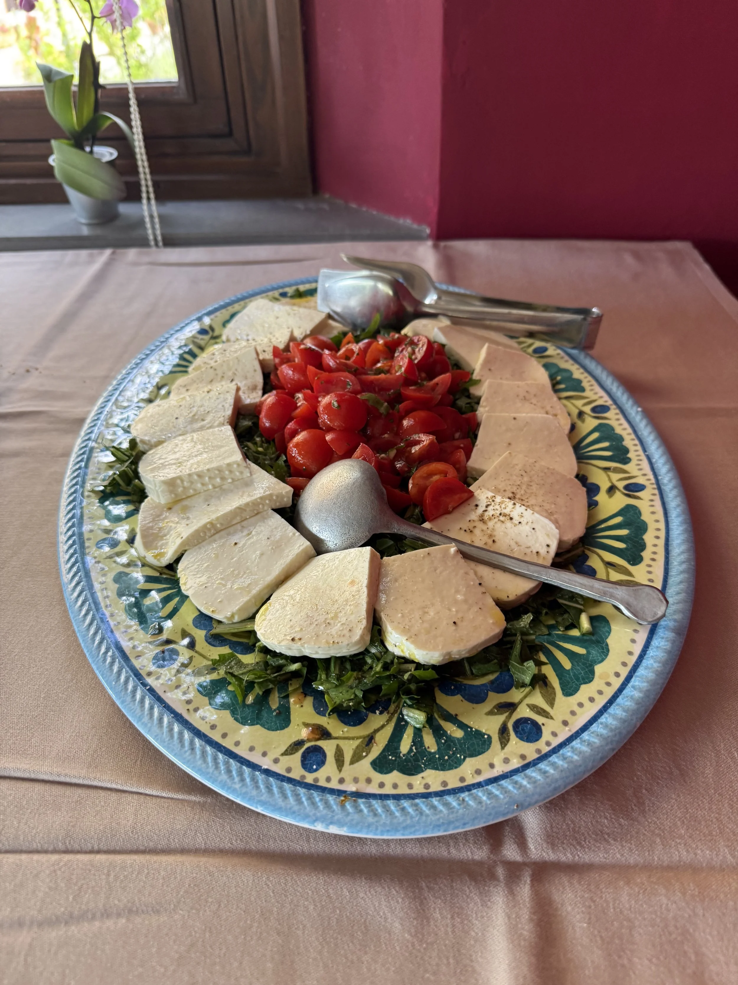 A decorative plate with sliced cheese, cherry tomatoes, and sliced deli meats on a table with a pink tablecloth, near a window with a potted orchid plant.