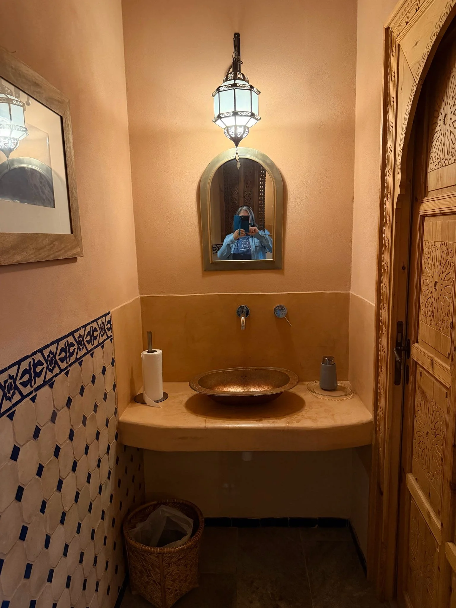 A bathroom with a small sink, a mirror, a hanging lantern light fixture, and decorative wooden doors. There is a paper towel holder, a trash can, and some small bottles on the counter.