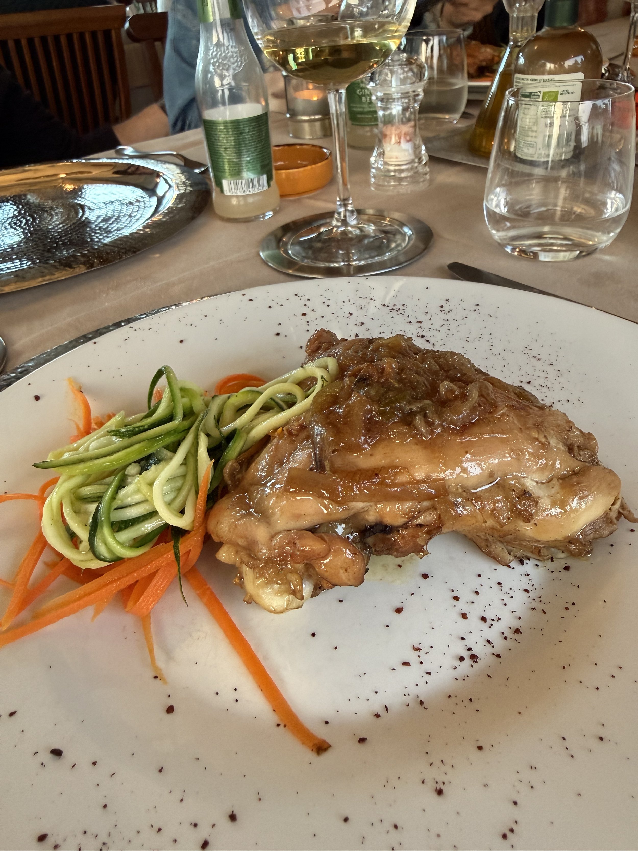 A white plate with cooked chicken topped with sauce, grilled, with shredded carrots and zucchini noodles on the side. The table has glasses, bottles of water, and wine in the background, with a silver platter on the left.