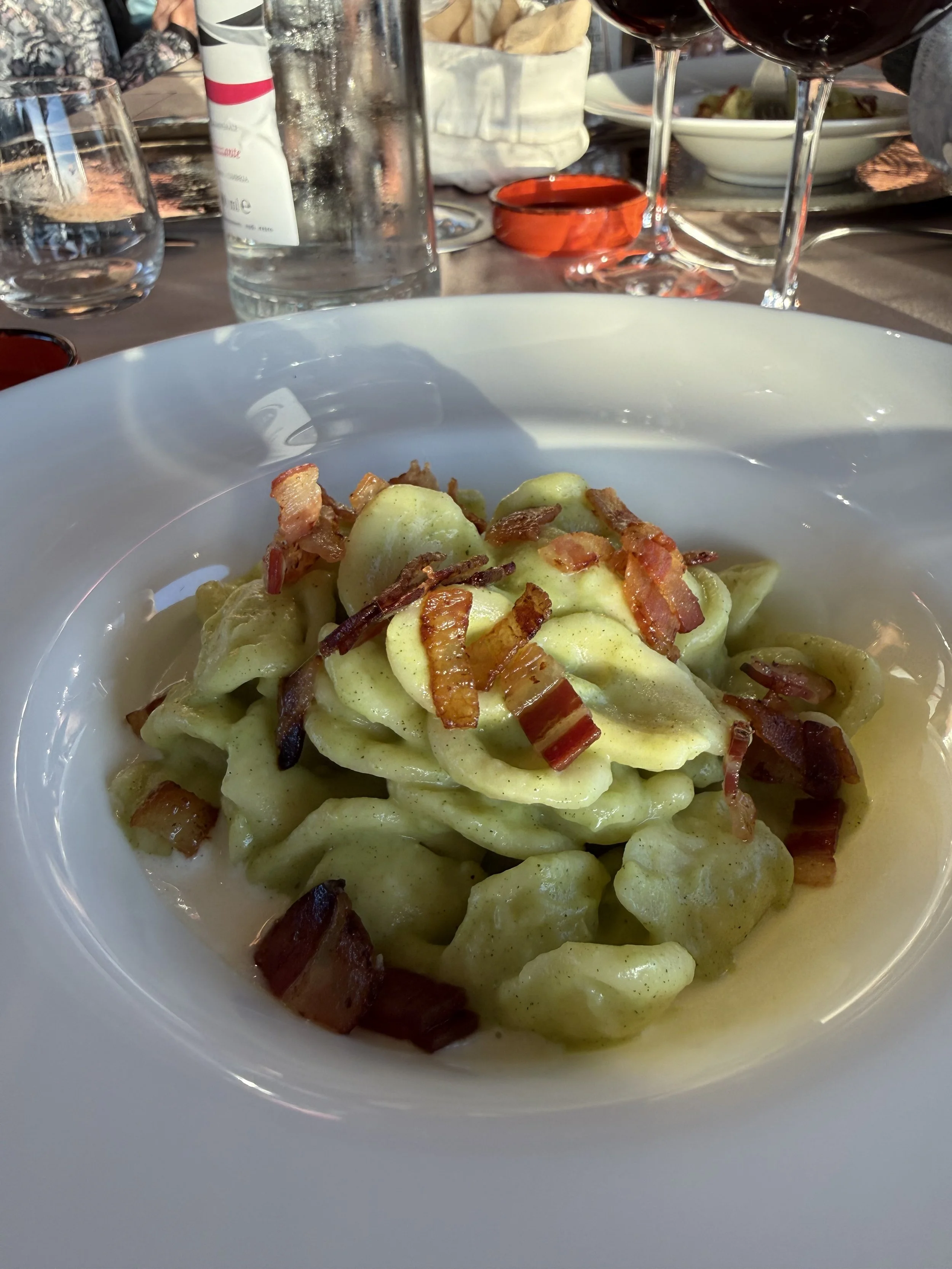 A bowl of pasta with bacon on top, set on a table with glasses of red wine, a water bottle, and other dishes in the background.