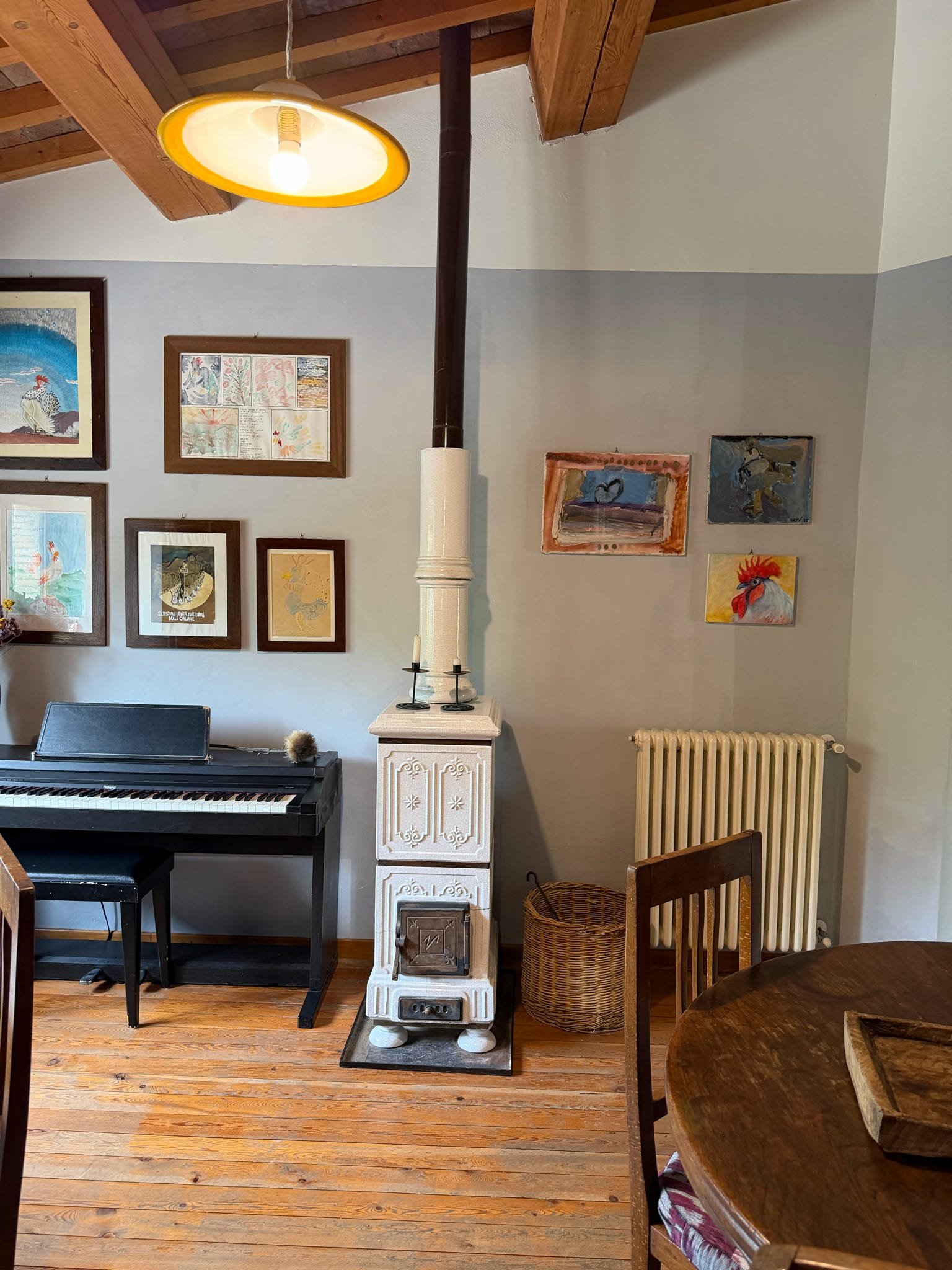 Image of a cozy living corner with a piano, a small decorative white stove, and wall art, with wooden floors, chairs, and a textured ceiling with wooden beams. A yellow pendant light hangs from the ceiling.