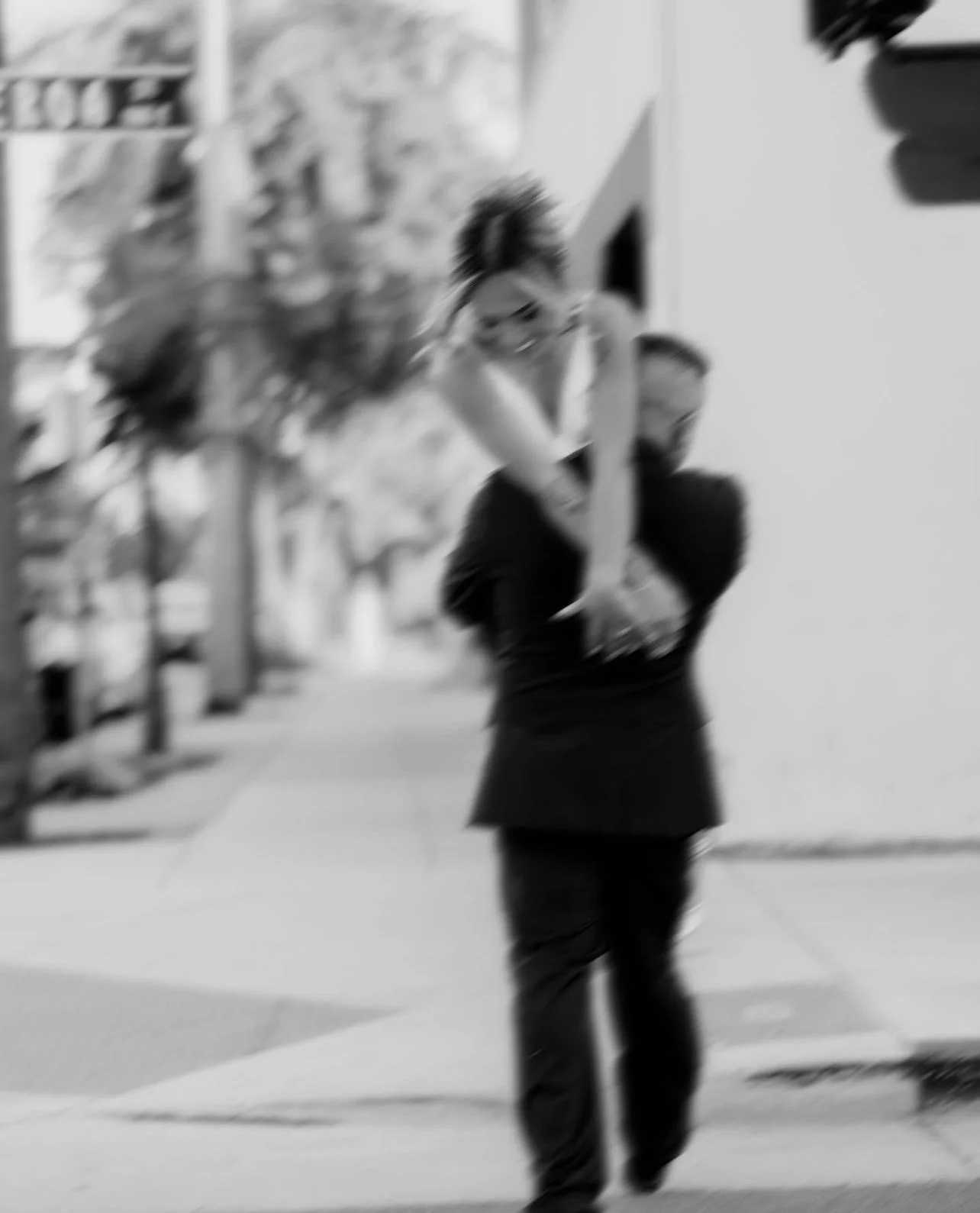 A blurred black and white photo of a person with short hair and glasses carrying a woman on their shoulders on a city sidewalk.