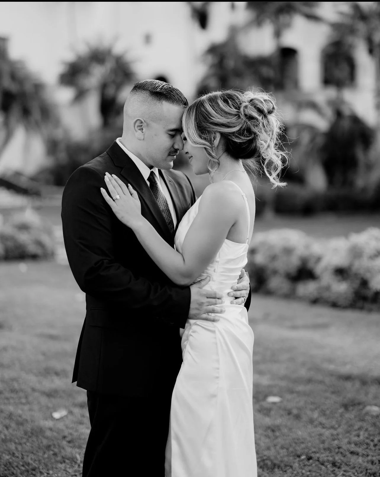 Black and white photo of a couple on their wedding day, embracing outdoors with heads touching, eyes closed, smiling gently, in a garden setting.
