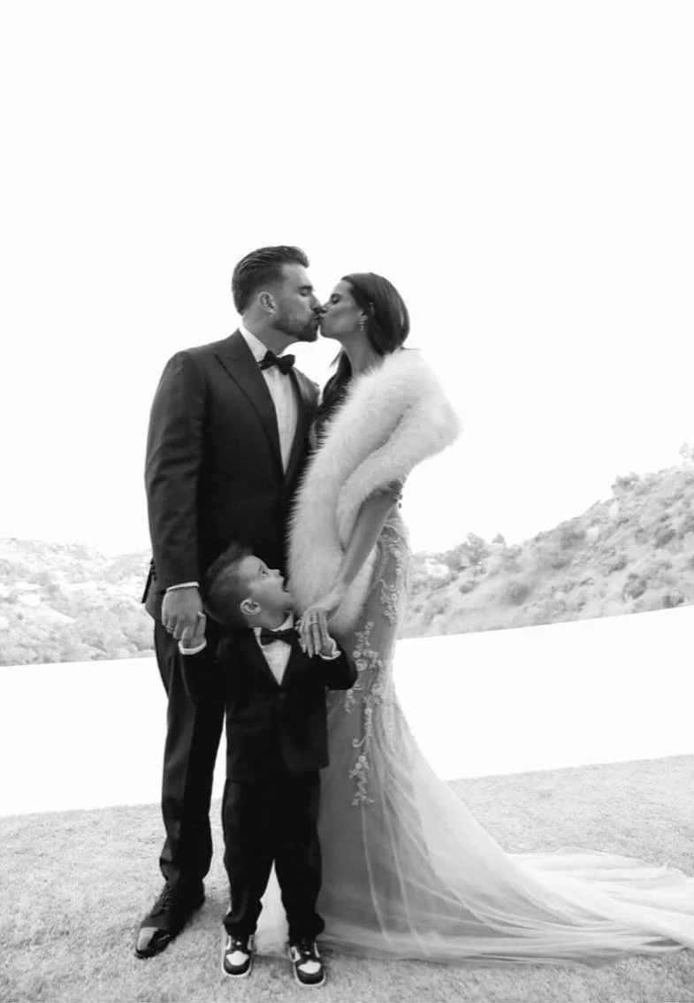 A black and white photo of a newlywed couple sharing a kiss, with a young boy looking up at them, outside in a snowy landscape.