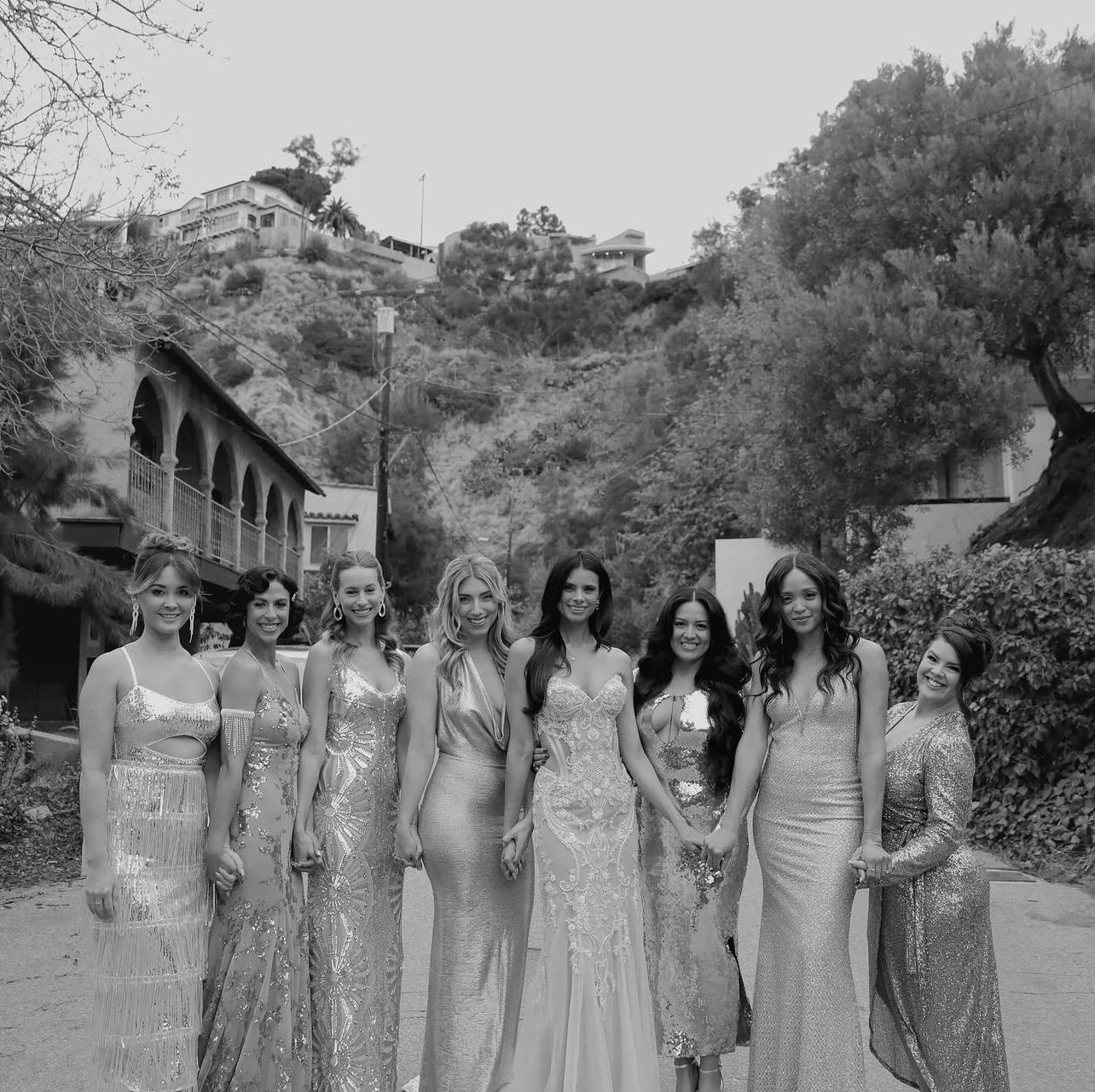 Group of eight women in evening gowns holding hands outdoors on a street, with hillside houses and trees in background.
