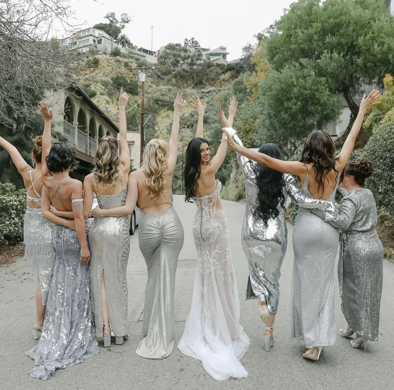 Group of women dressed in silver and white formal dresses, standing on a street with their backs to the camera, raising their arms in celebration.