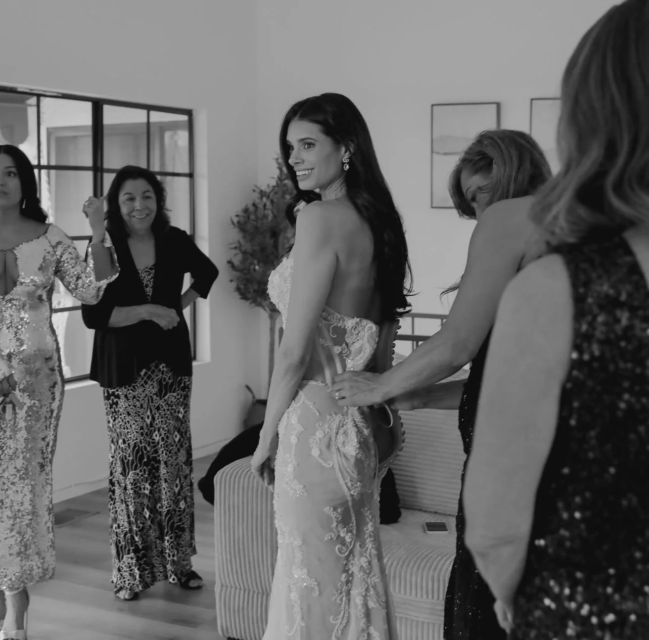 A bride in a wedding dress is being helped with her dress by a woman, and she's smiling at her. Several women surround them, some smiling and looking on, in a room with modern decor.