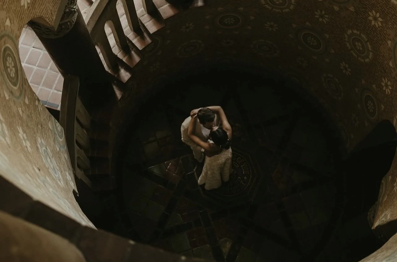 A couple standing on a dark tiled floor, seen from above, embracing and kissing.