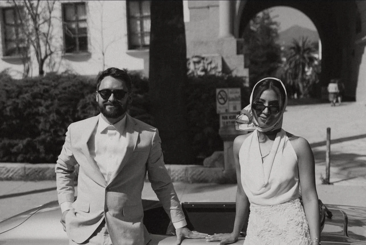 A black and white photo of a man and woman standing outside in front of a vintage car, dressed in formal attire, both wearing sunglasses. The background features trees, a building, and an archway.