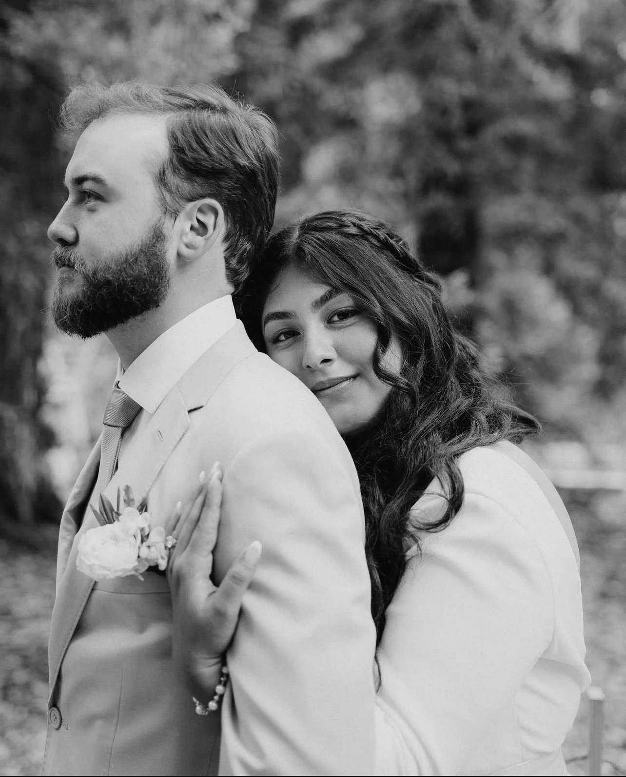 A black and white photograph of a couple, with the woman embracing the man from behind, resting her chin on his shoulder, outdoors with trees in the background.