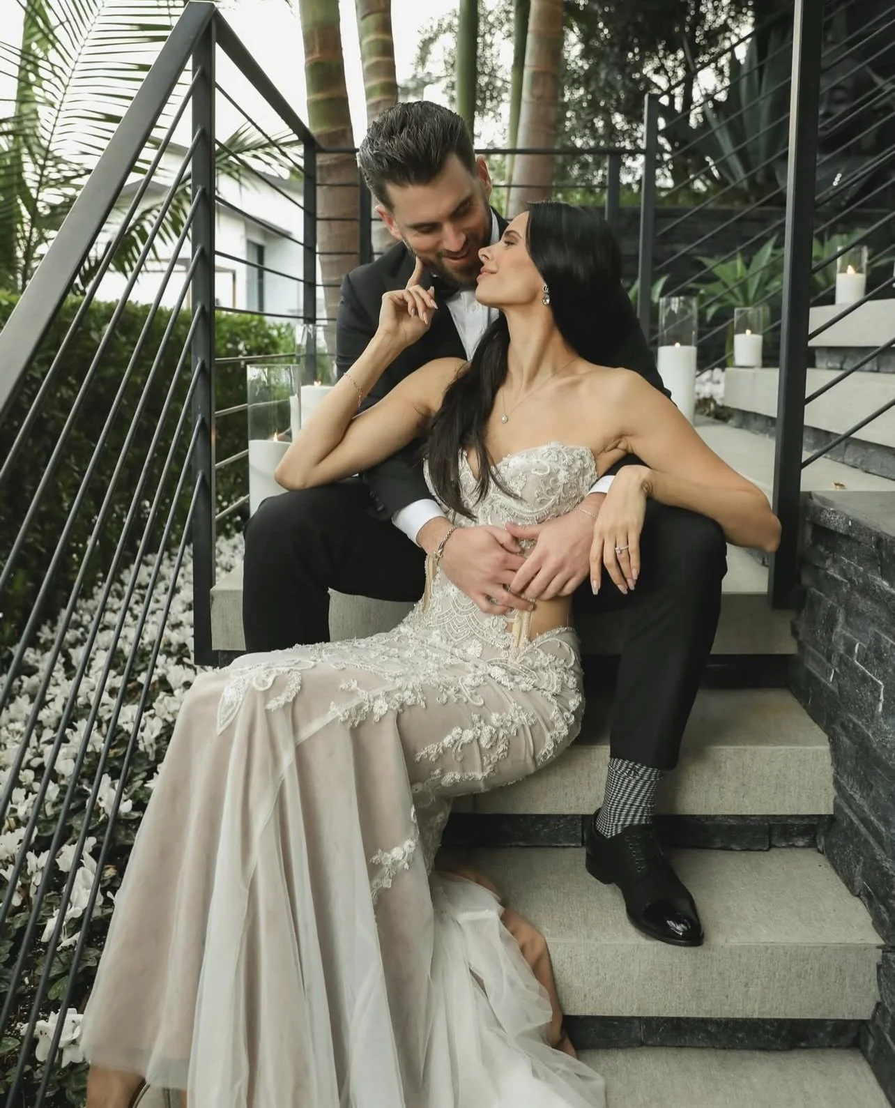 A couple dressed in wedding attire sitting on outdoor stairs, with the woman in a white lace wedding dress and the man in a tuxedo. They are close, exchanging a tender moment, surrounded by candles and greenery.