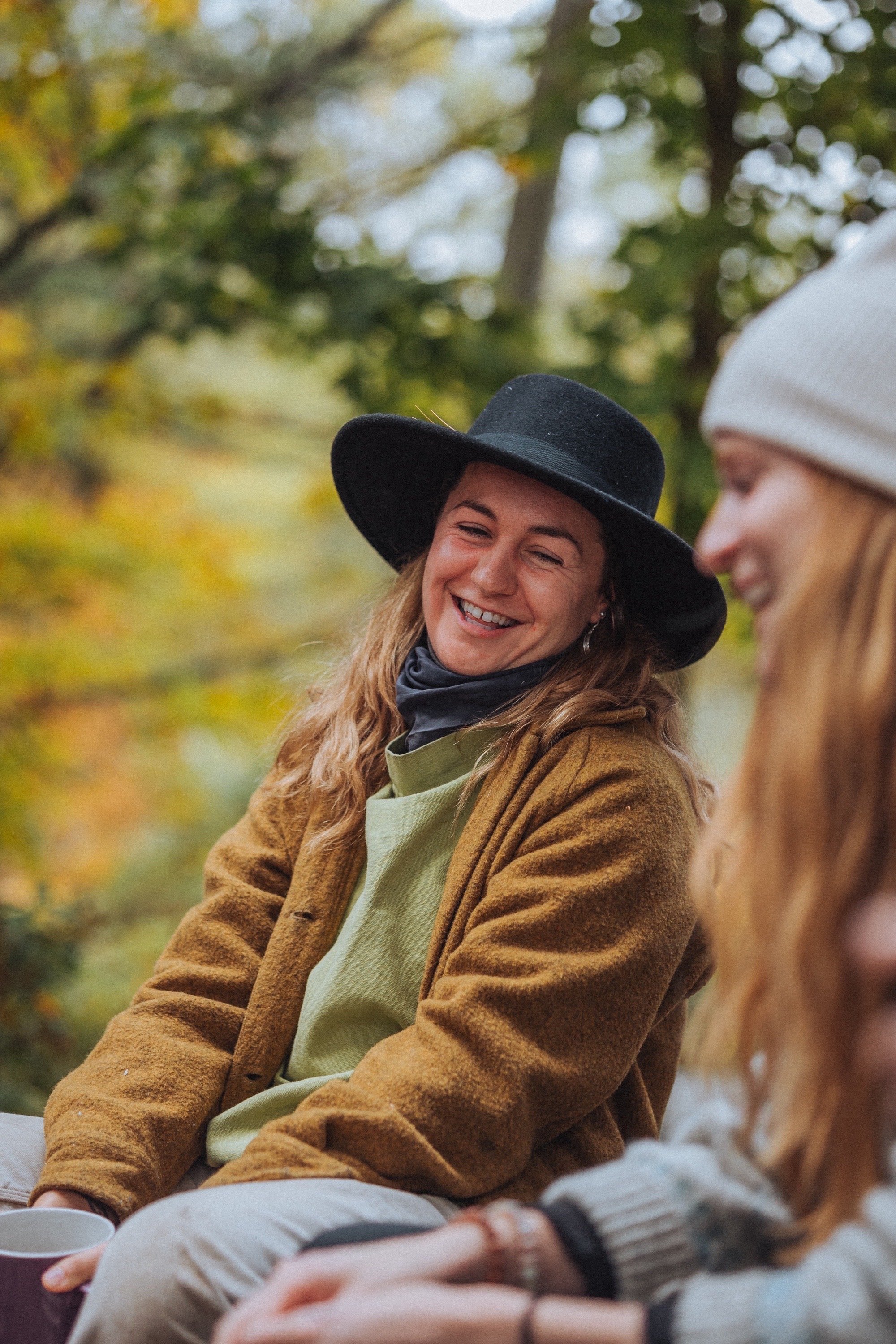Two people sitting outdoors, one wearing a black hat and brown jacket, smiling, with trees in the background.
