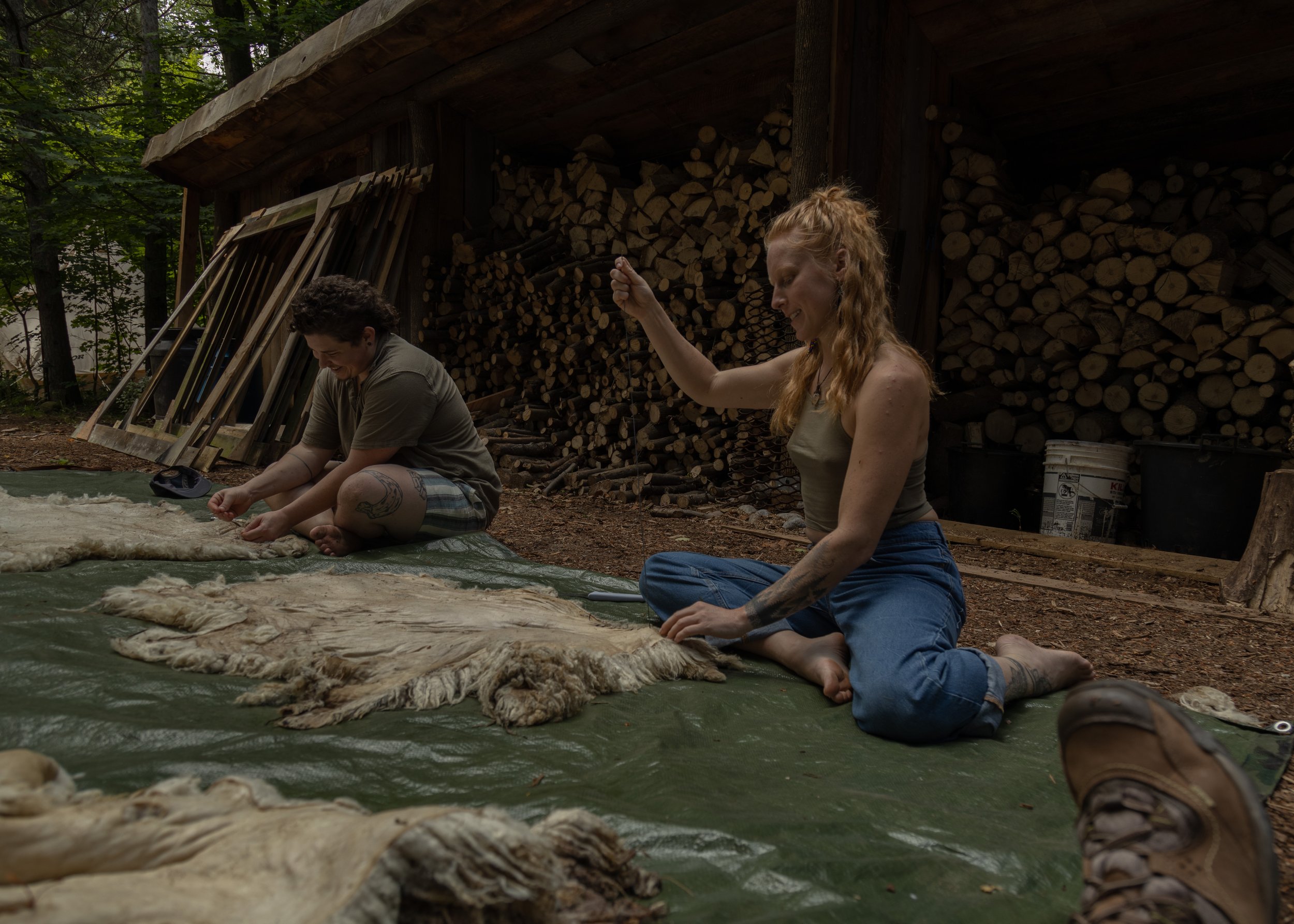 Two people sitting on the ground, working with wool or hides in a wooded area near a stack of firewood.