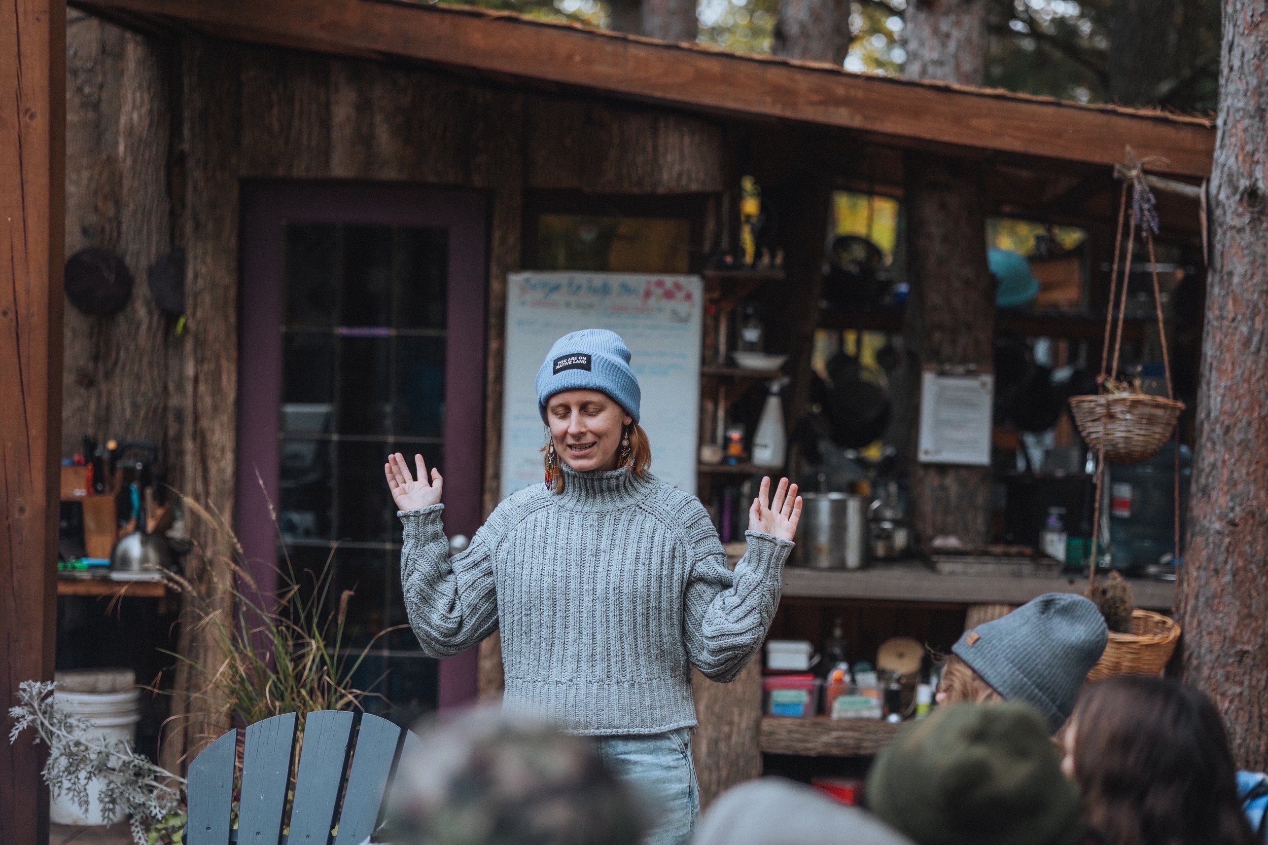 Person speaking outdoors in front of a rustic building, wearing a gray knit sweater and a light blue beanie, with hands raised. Audience members visible at the bottom.