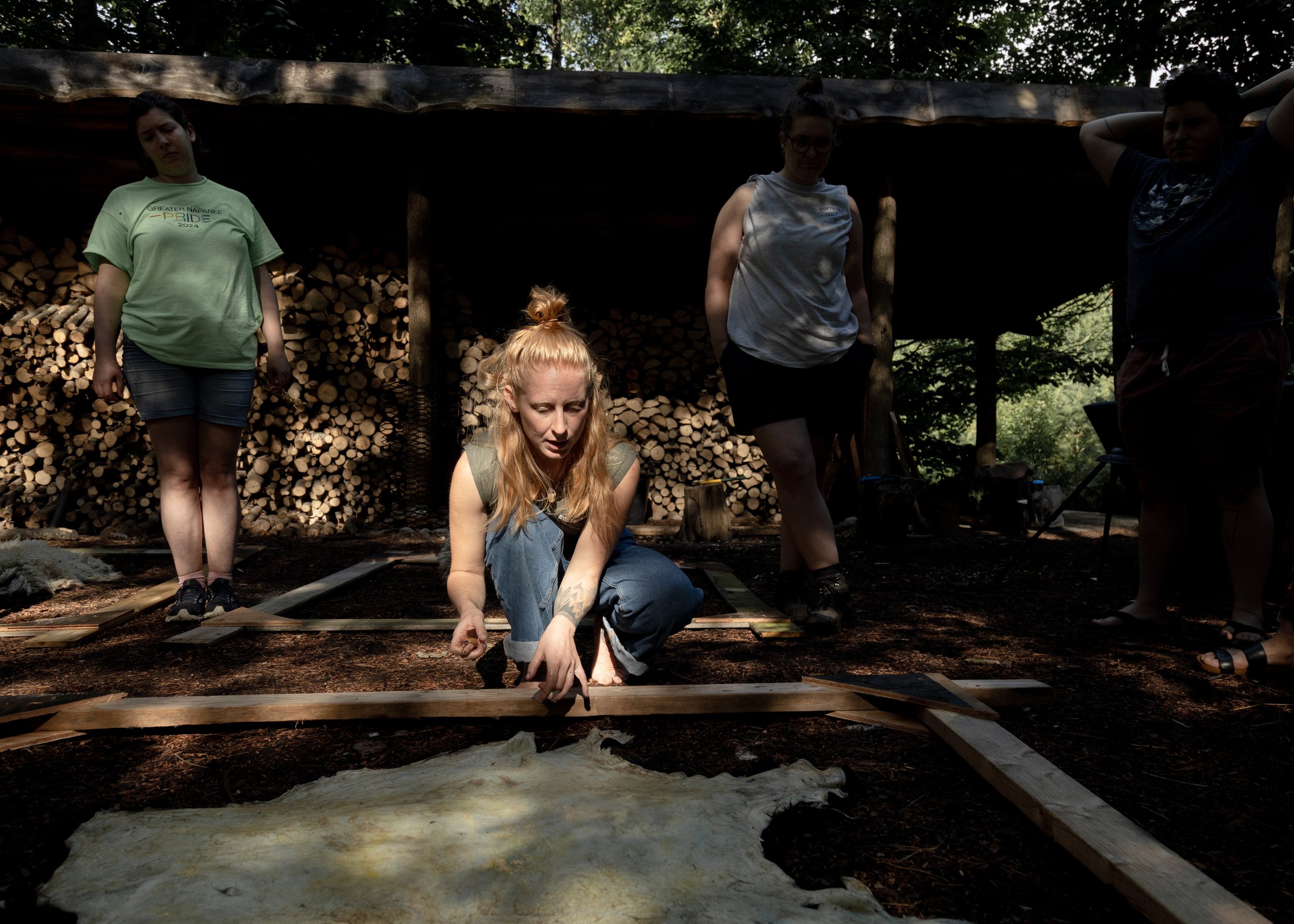A person kneeling and working on animal hide outdoors, surrounded by three others standing, with a backdrop of stacked firewood.