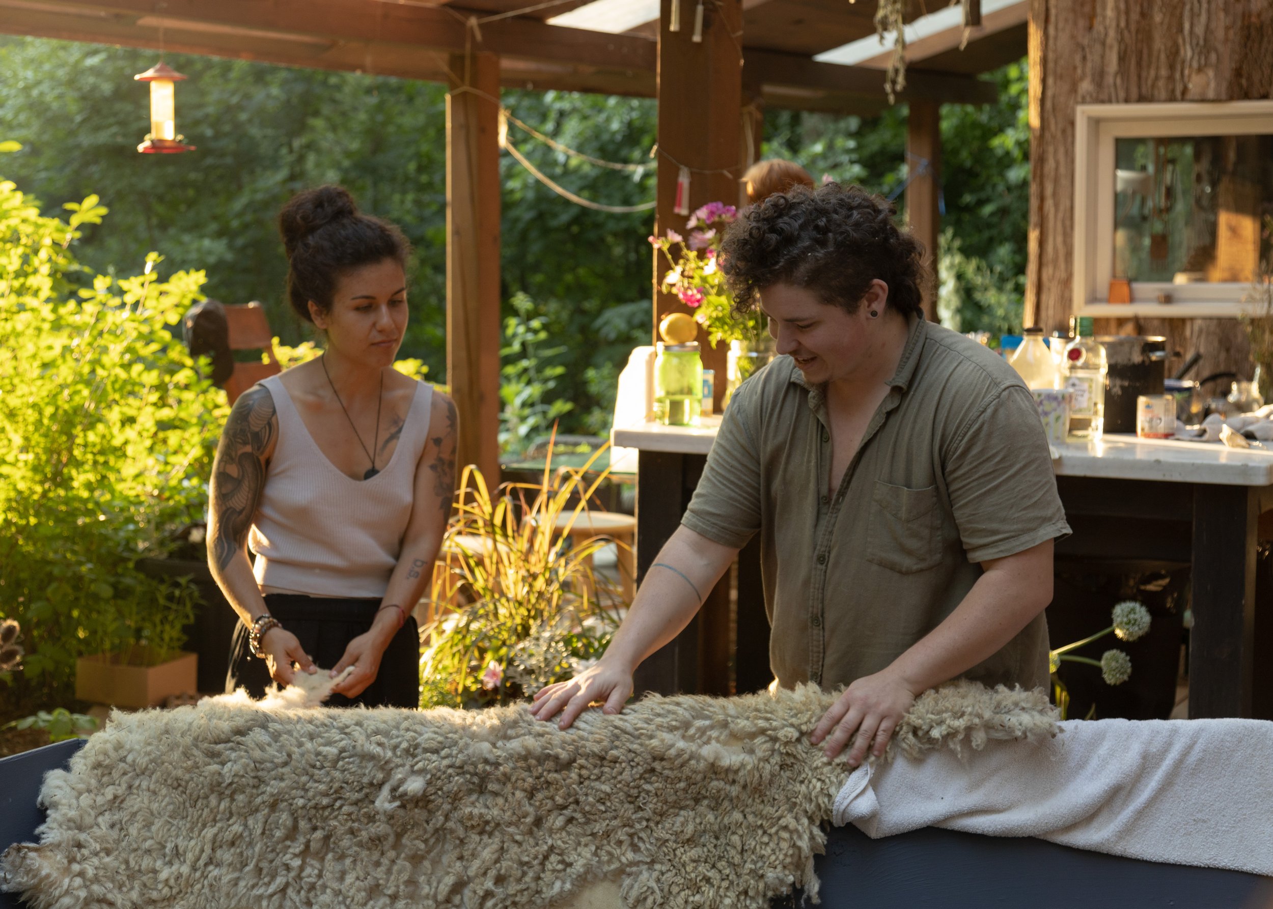 Two people working with fleece outdoors, surrounded by plants and sunlight, in a rustic setting.