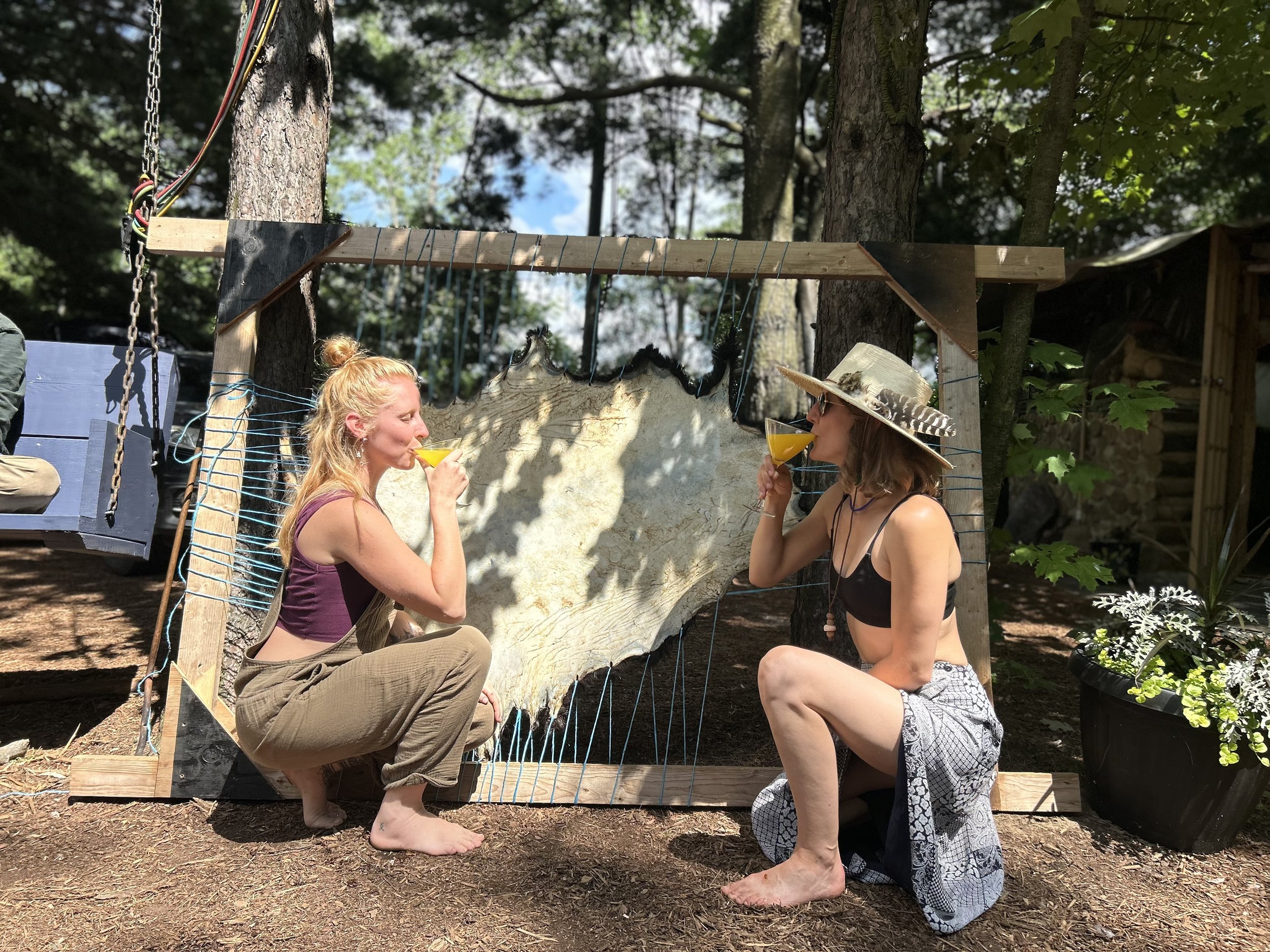 Two women squatting outdoors near a wooden frame with a hide or large cloth stretched across it. One woman on the left has blonde hair and is holding a yellow drink, wearing brown overalls and a purple top. The woman on the right wears a hat, black t
