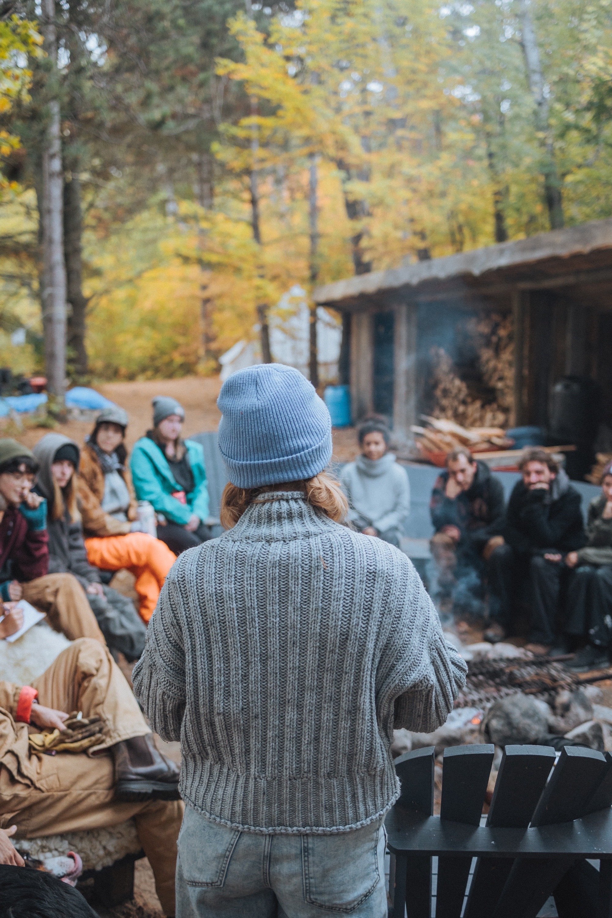 A group of people sitting outdoors in a circle, having a discussion. The person in focus is wearing a gray sweater and blue hat. Other participants are dressed warmly and surrounded by trees with autumn foliage.