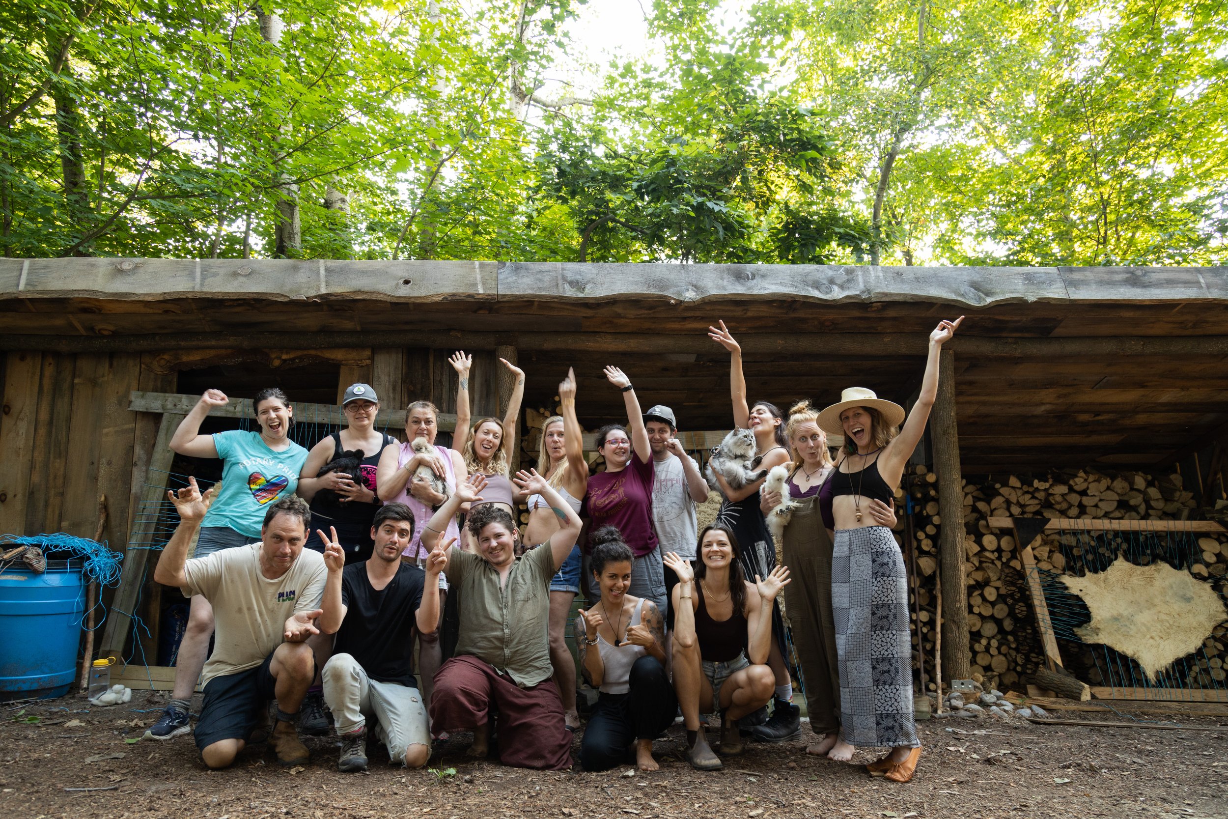 A group of people posing outdoors in front of a wooden structure in a forest. Some individuals are holding small animals. They appear cheerful and are making playful gestures.