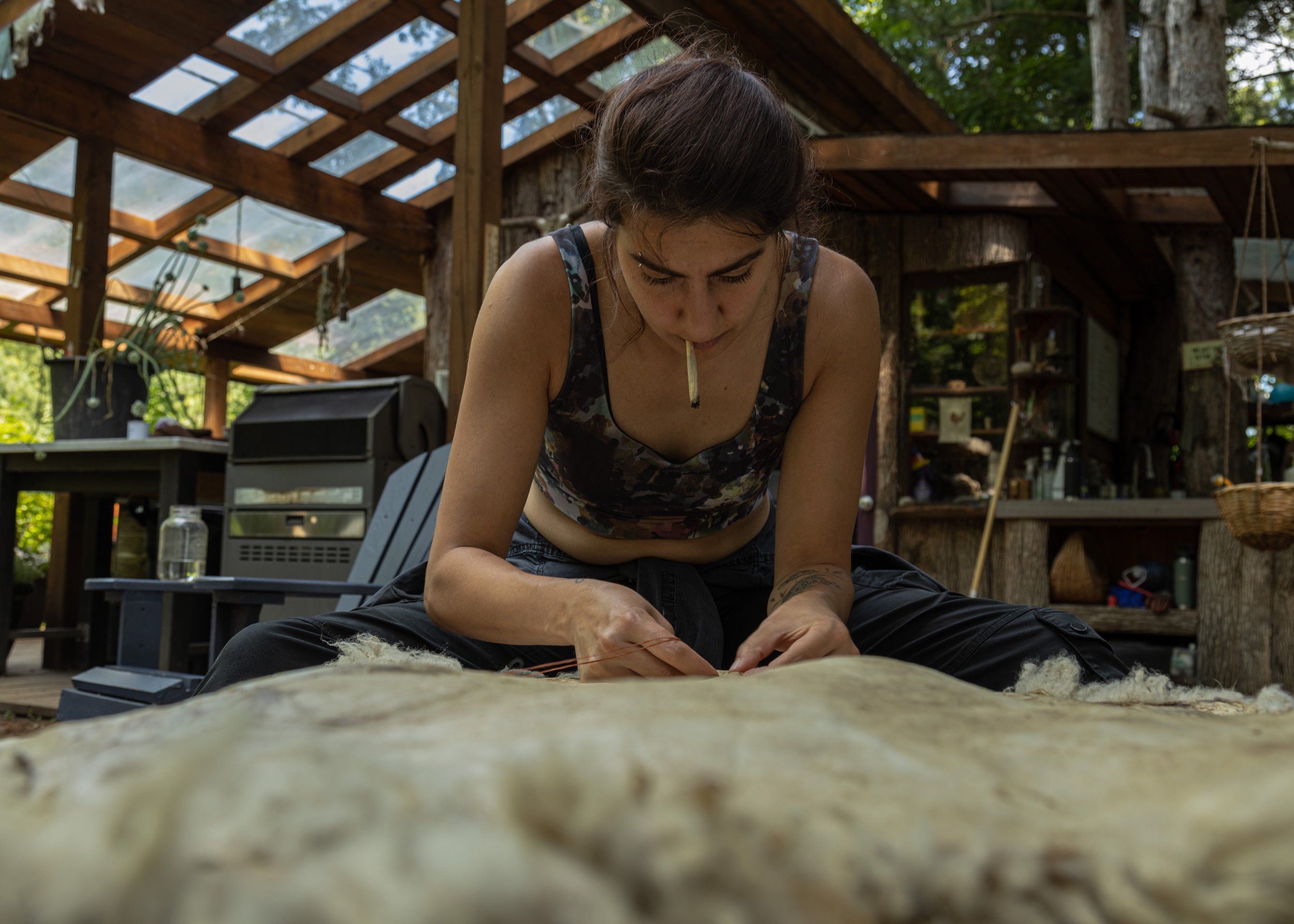 A person engaged in a craft or repair work inside a rustic greenhouse or cabin, with a rolled paper in their mouth. They are focused on sewing or assembling something on a wooden table. The setting includes wooden beams, plants, and outdoor equipment