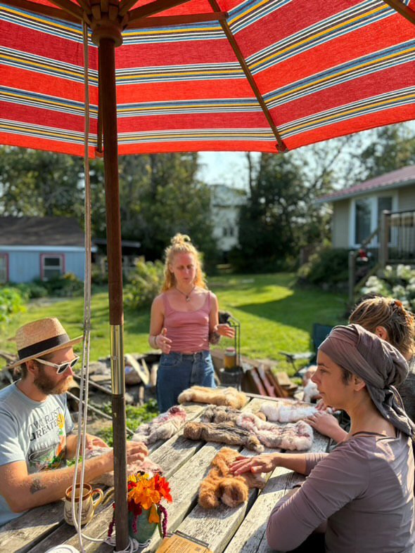 A group of people sitting around a wooden table outdoors under a red striped umbrella, engaging in a workshop or casual meeting. Various soft materials or fabrics are spread out on the table. The setting is a sunny backyard with grass and trees in th