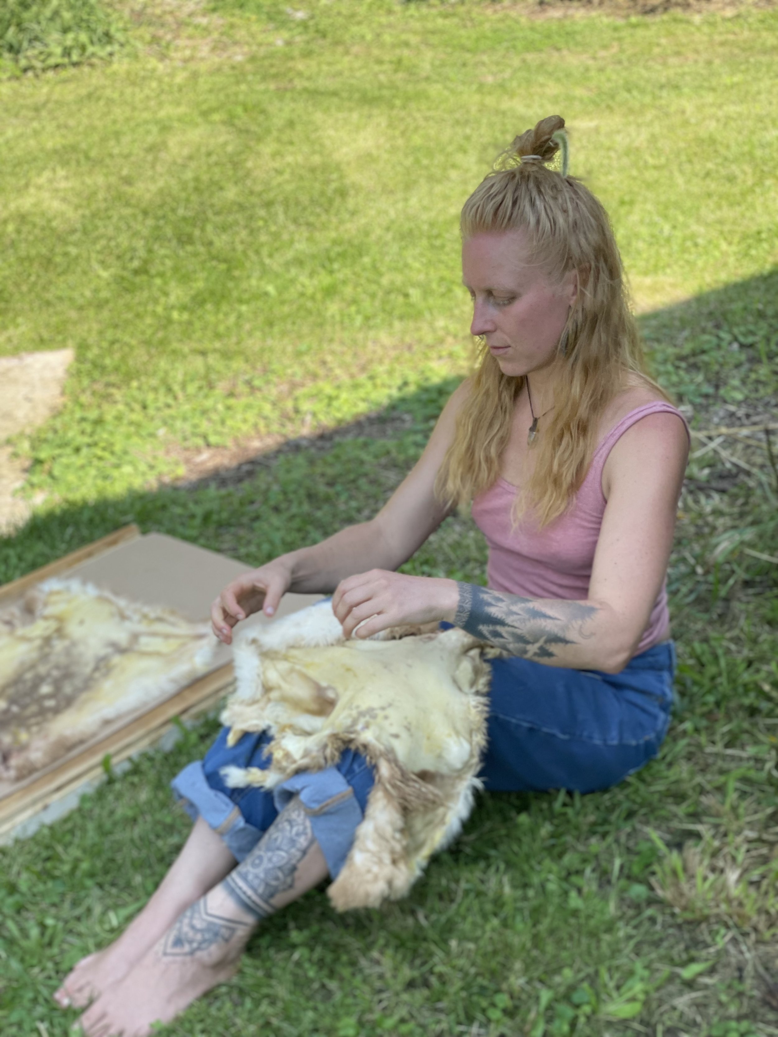 Person with tattoos sitting on grass, working on animal hide