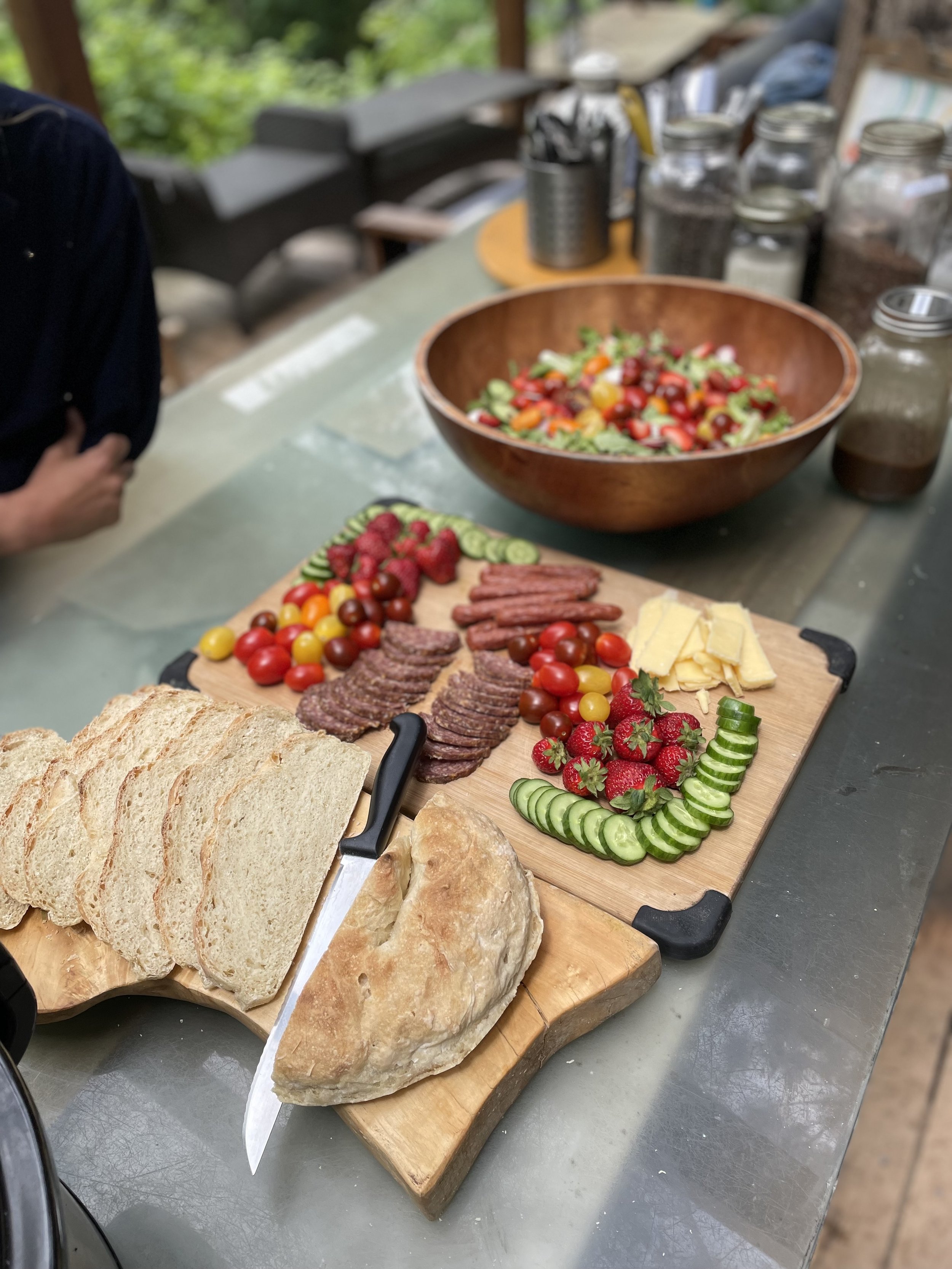 Charcuterie board with bread, salami, cheeses, strawberries, cucumbers, cherry tomatoes, and a salad in a wooden bowl.