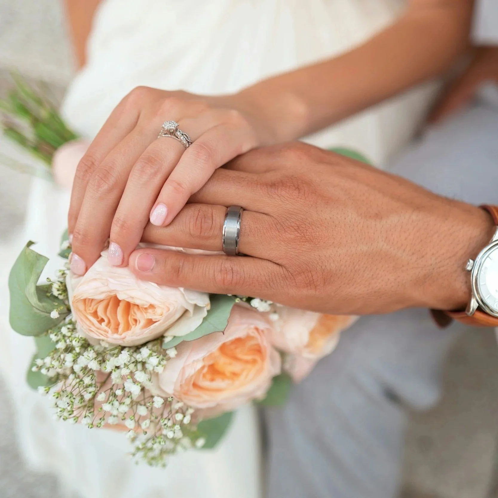 Close-up of newlywed couple's hands with wedding rings, resting on a bouquet of peach and white roses and baby's breath.