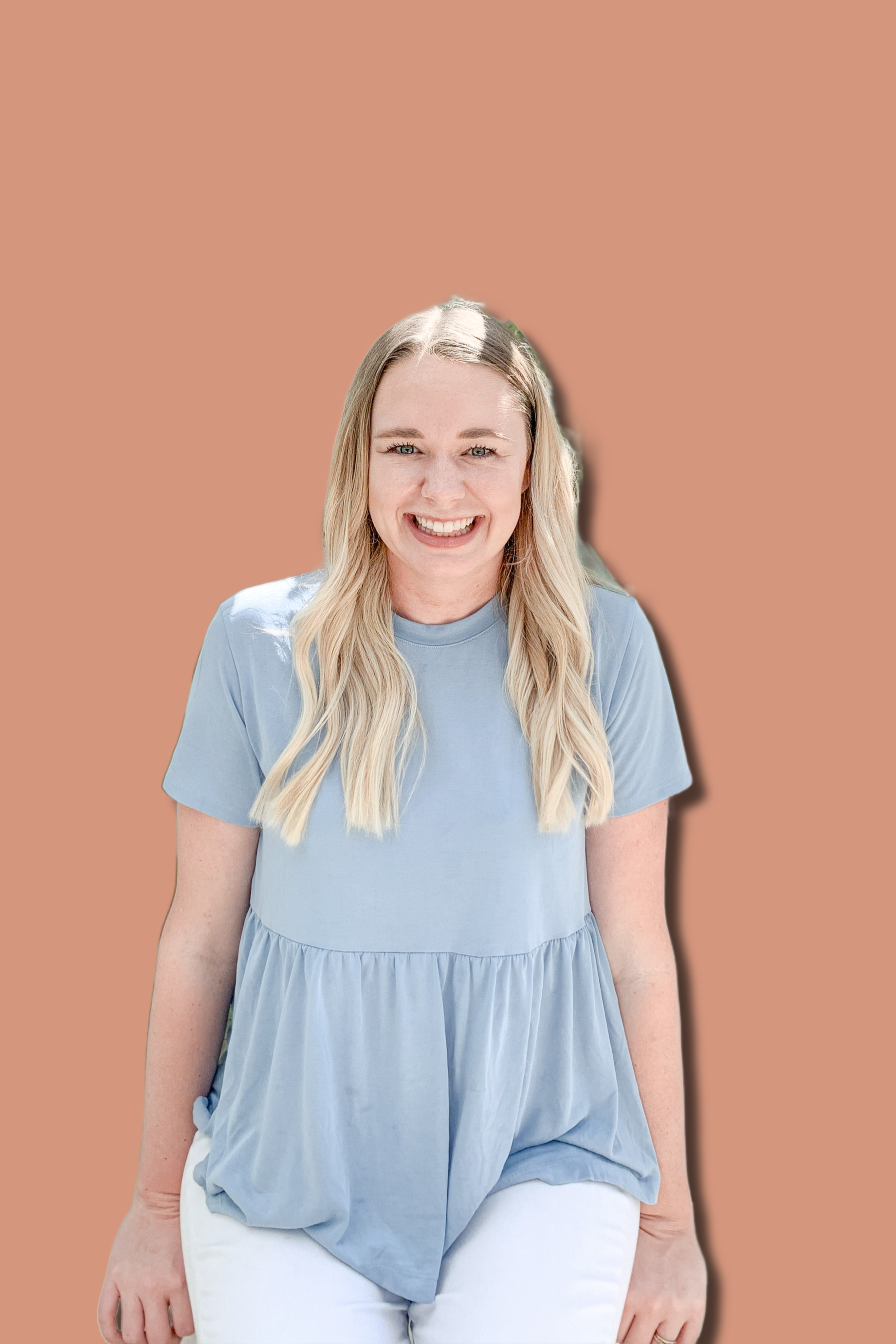 A woman with long blonde hair smiling, wearing a light blue top and white pants, standing against a solid light brown background.