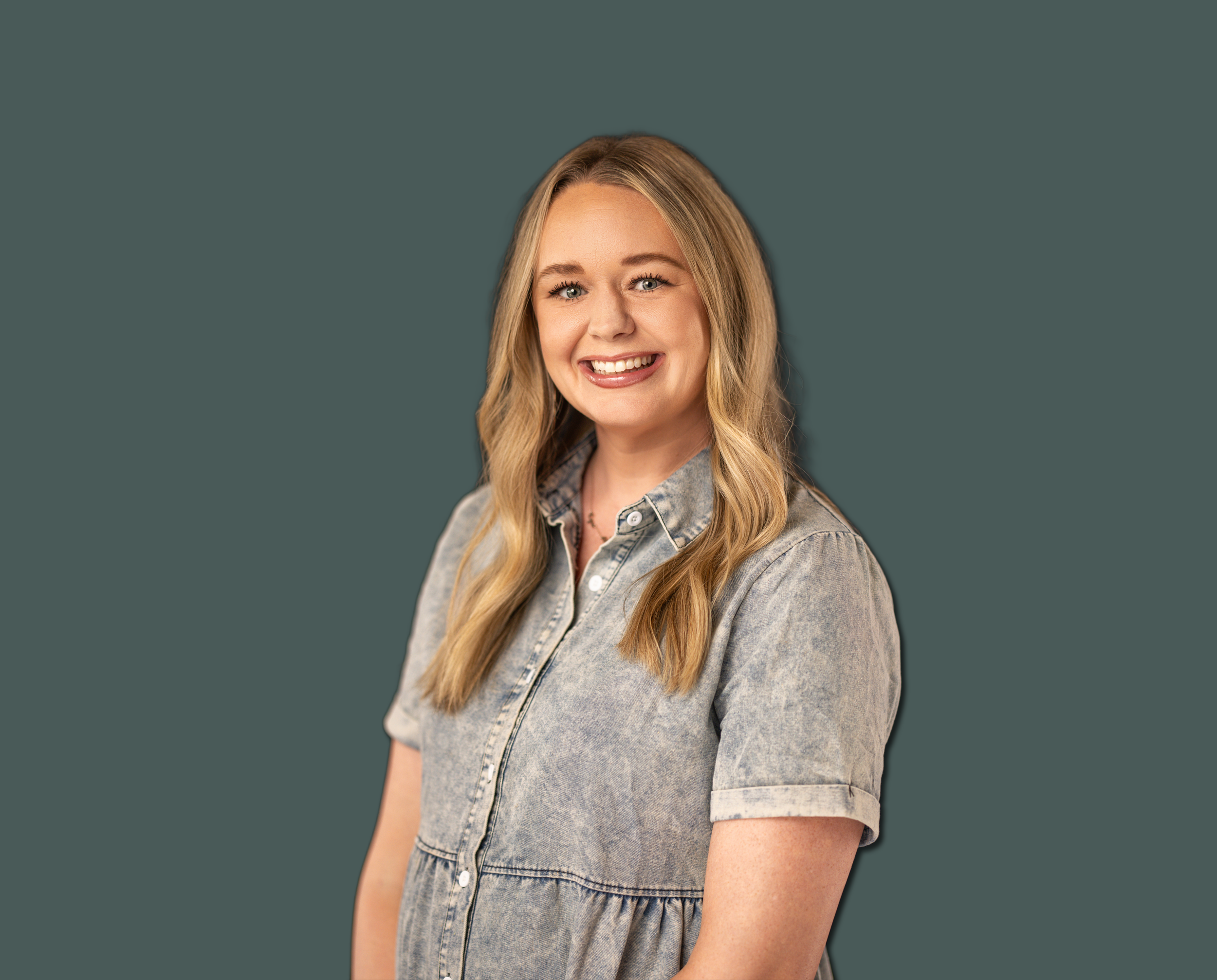 A young woman with blonde hair smiling, wearing a denim dress, standing against a plain dark teal background.