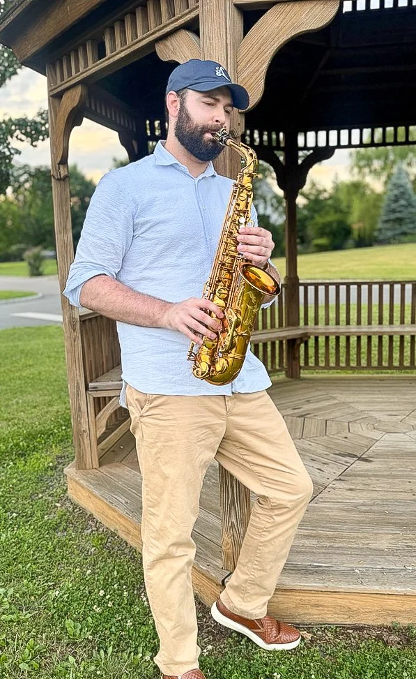 A man with a beard, wearing a cap, light blue shirt, khaki pants, and brown shoes, playing a gold-colored saxophone outdoors near a wooden gazebo with greenery in the background.