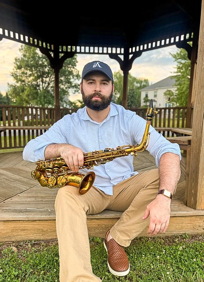 A man sitting on a wooden porch holding a gold saxophone, wearing a light blue shirt, tan pants, brown shoes, a black watch, and a dark blue cap with a white logo, with trees and a house in the background.