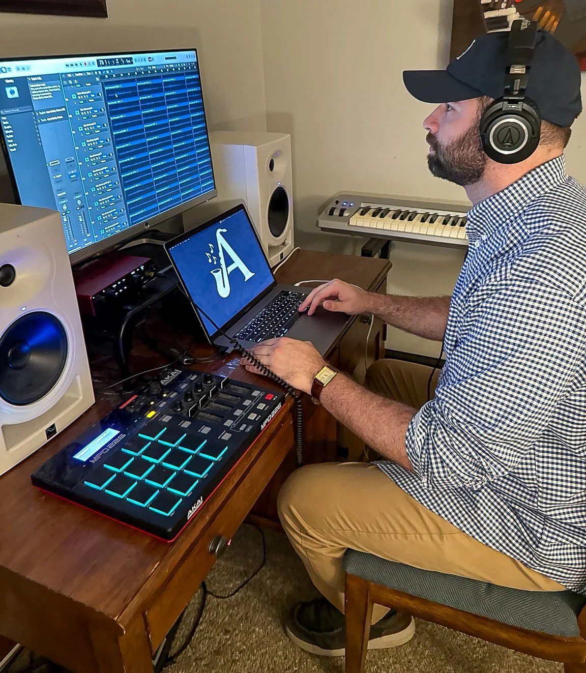 Man wearing headphones and a cap working on music production in a home studio with a computer, MIDI controller, speakers, and a keyboard.