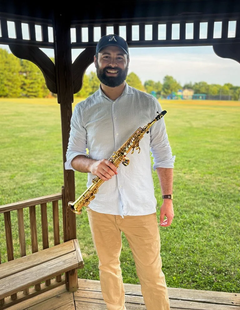 A man with a beard and a cap is standing outside under a wooden gazebo, holding a gold-colored saxophone, smiling at the camera.