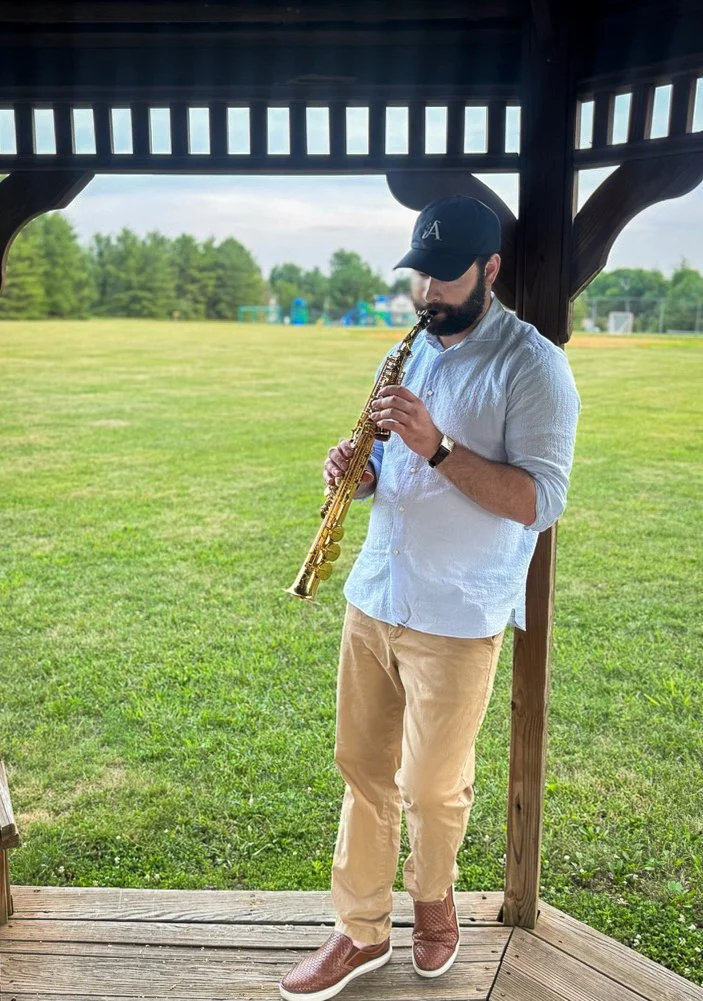 Man in light blue shirt, beige pants, brown shoes, and dark baseball cap playing a saxophone under a wooden pavilion in a park with green grass and trees.