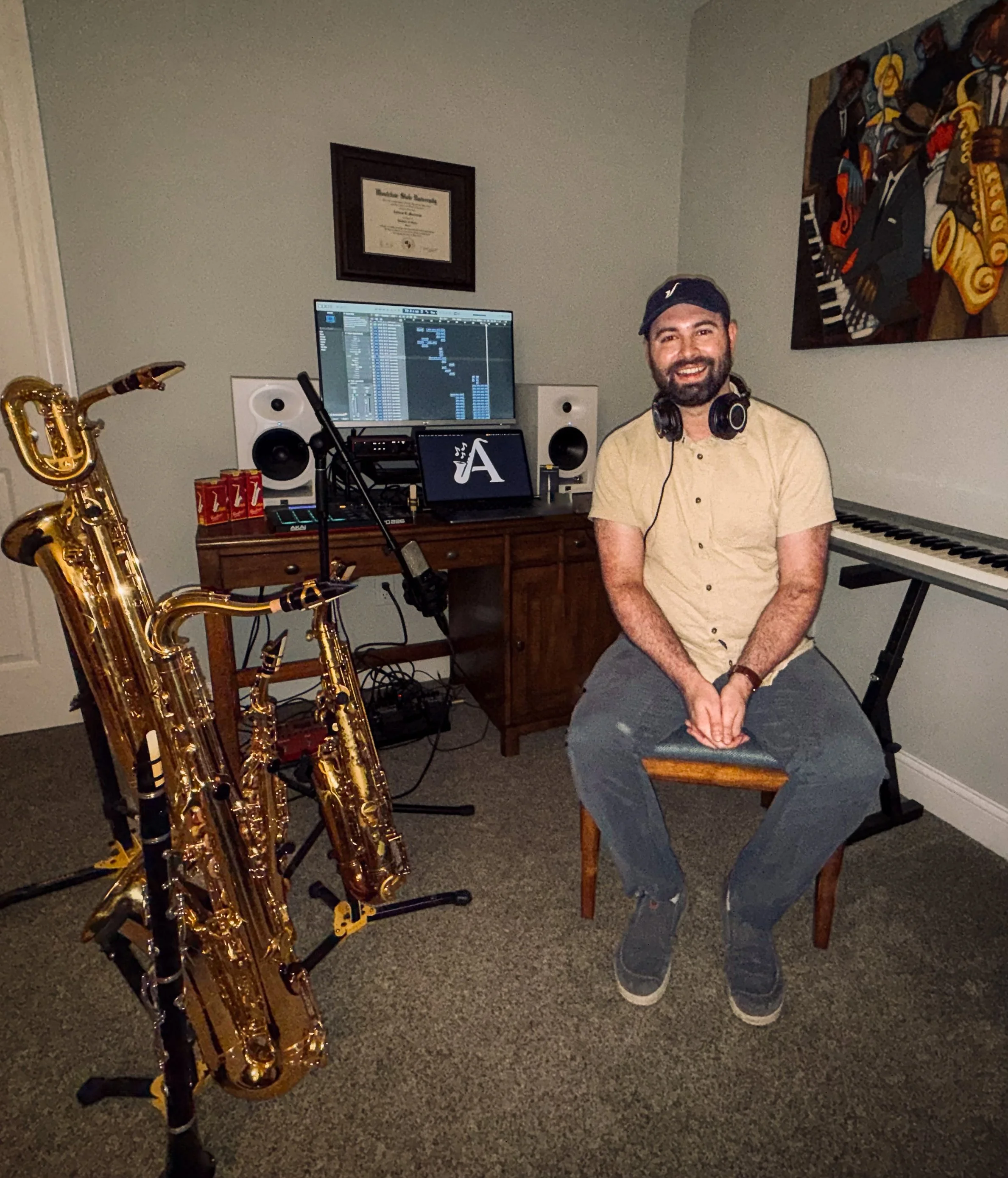 A man sitting on a wooden chair in a room with music equipment, including a keyboard, a computer screen, speakers, and saxophones. The man is smiling, wearing a baseball cap, headphones around his neck, a beige shirt, and gray pants. There is a framed certificate on the wall and a colorful painting to the right.