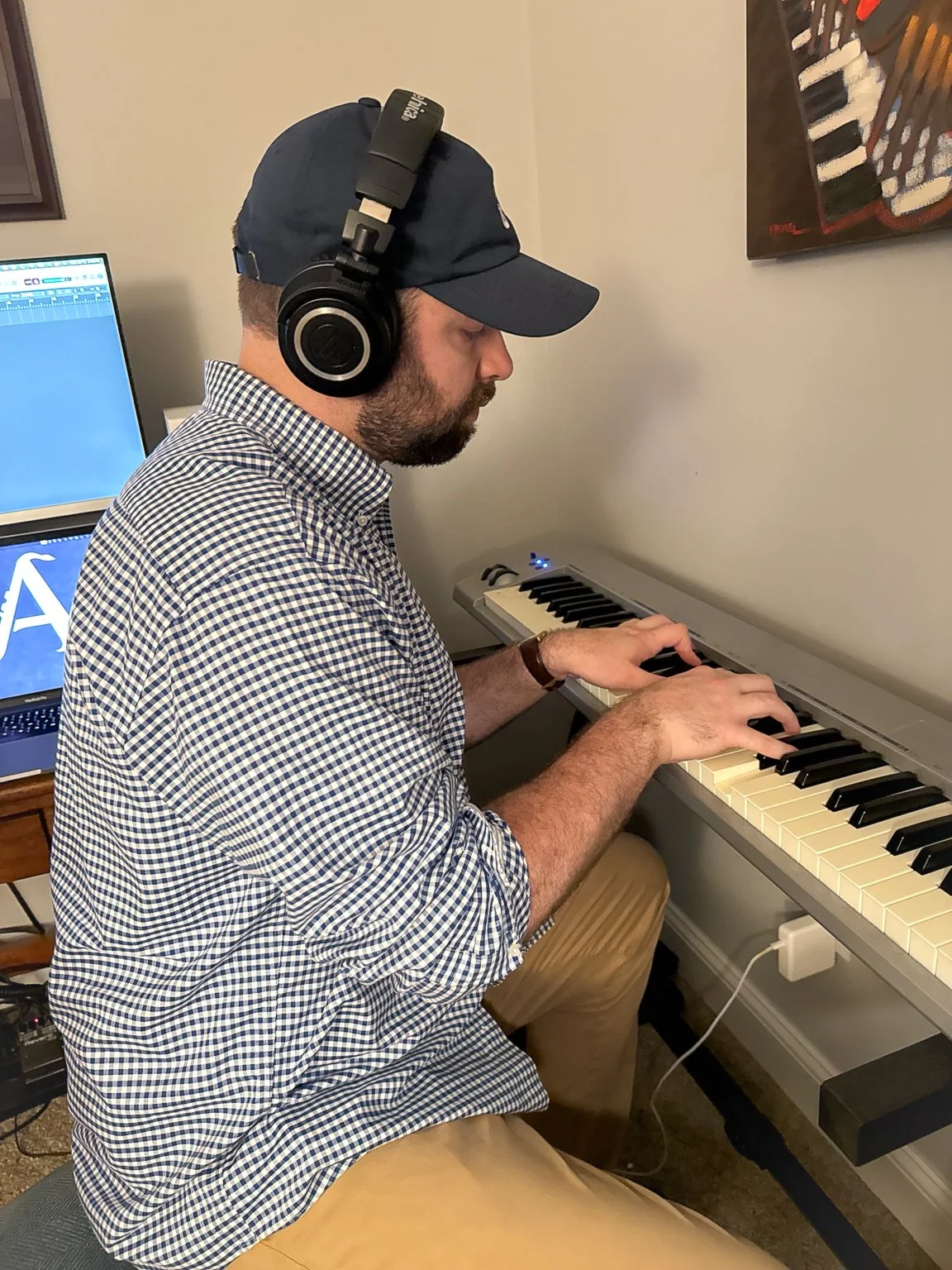 A man wearing a navy baseball cap and checkered shirt playing an electronic piano keyboard in a room, with a computer monitor visible in the background.