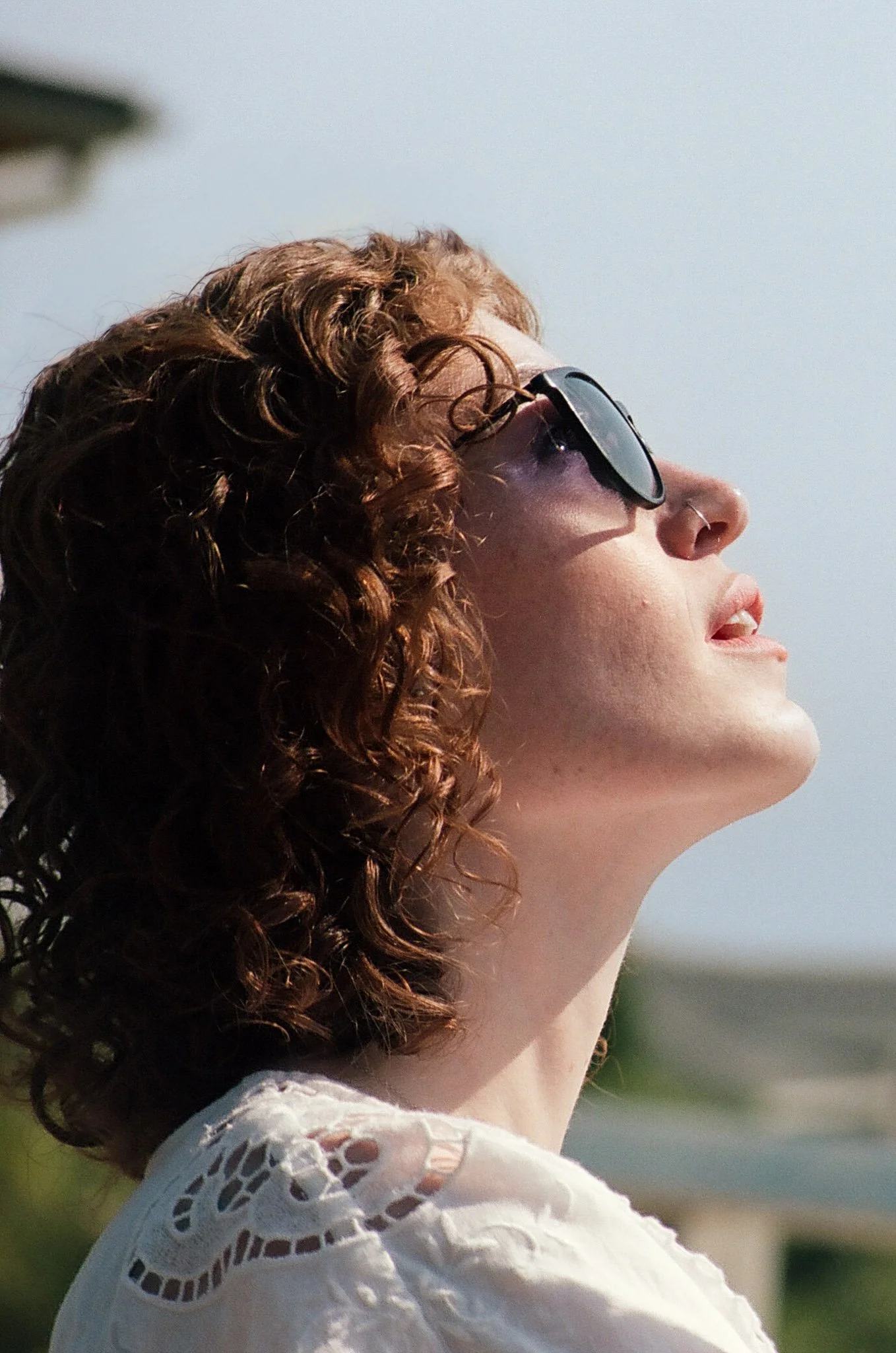 A woman with curly brown hair wearing sunglasses, looking upwards against a clear sky.